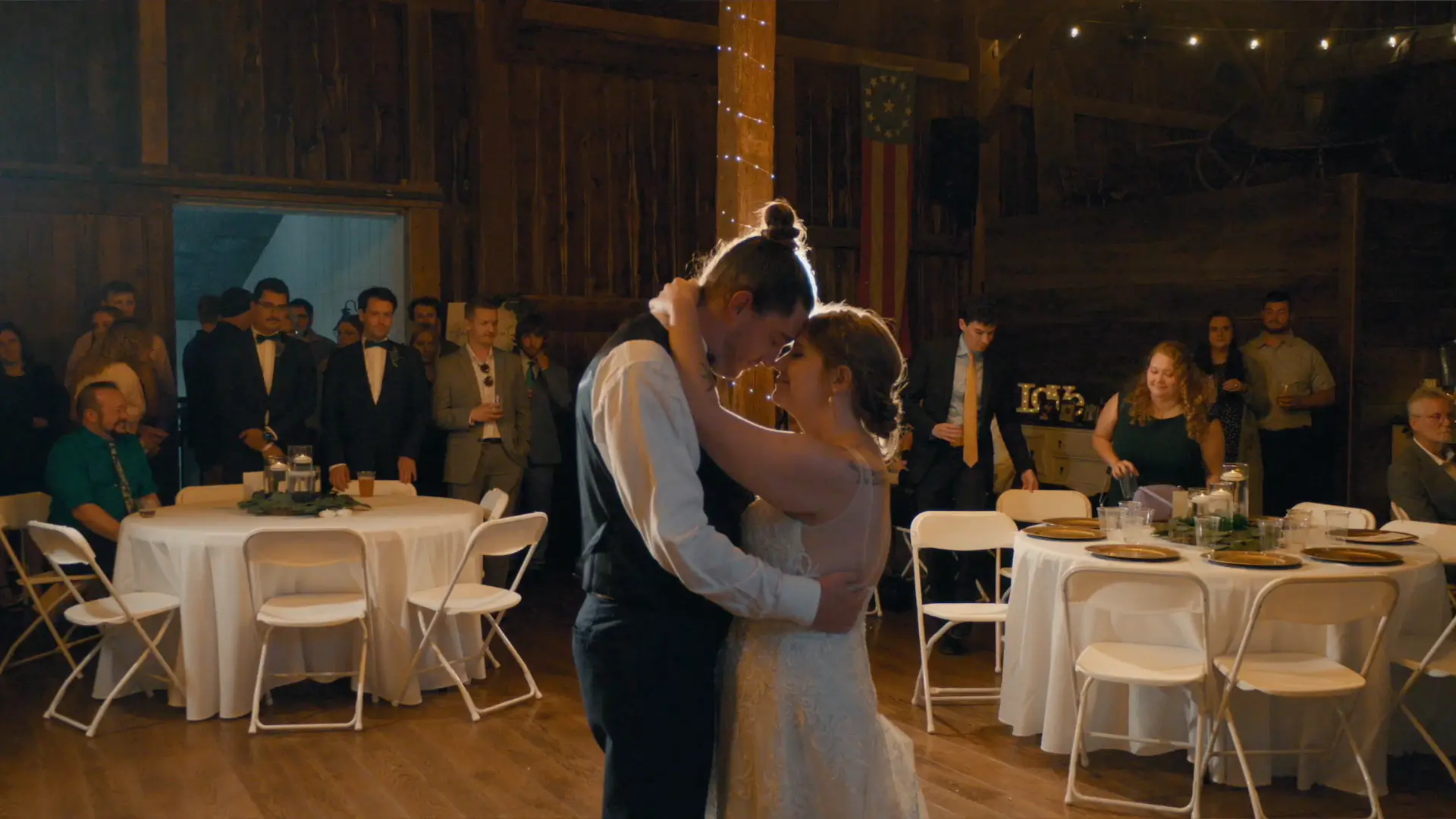 A newlywed couple shares a romantic first dance in a warmly lit barn venue, with guests seated and standing around watching. White chairs and tables with simple decor are visible, and string lights add a cozy ambiance.