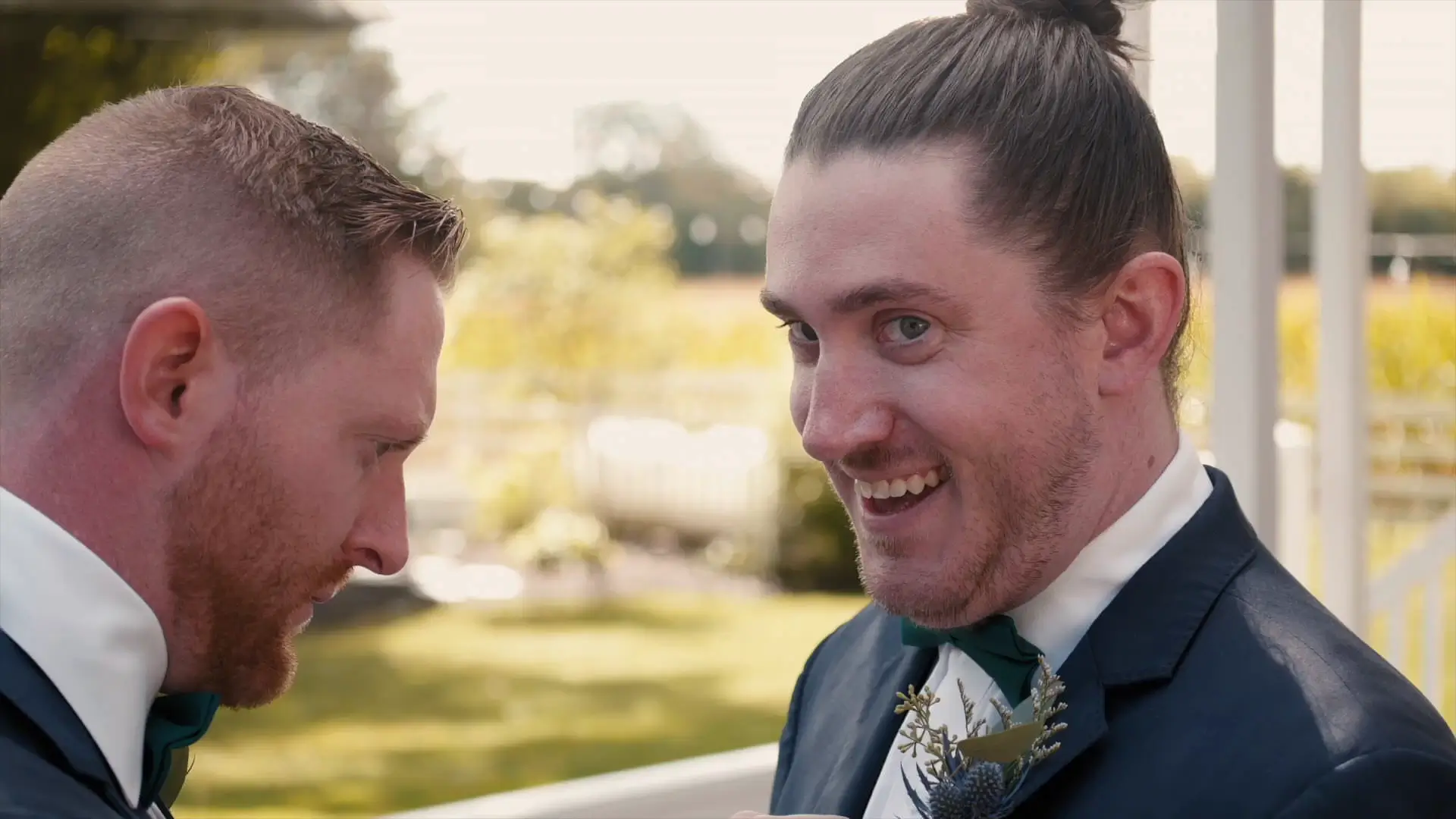 The groom shares a goofy smile with the camera as a groomsman looks down, fixing the groom's tuxedo before the ceremony at Barn1888 Events. It is a bright, sunny Michigan day with blurred greenery in the background, creating a joyful and light atmosphere.