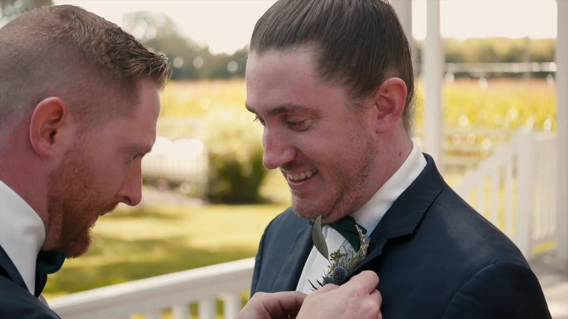 A groomsman adjusts the boutonniere on the groom's suit lapel, both smiling, on a sunny day at an outdoor venue with greenery in the background, as captured by their wedding videographer.