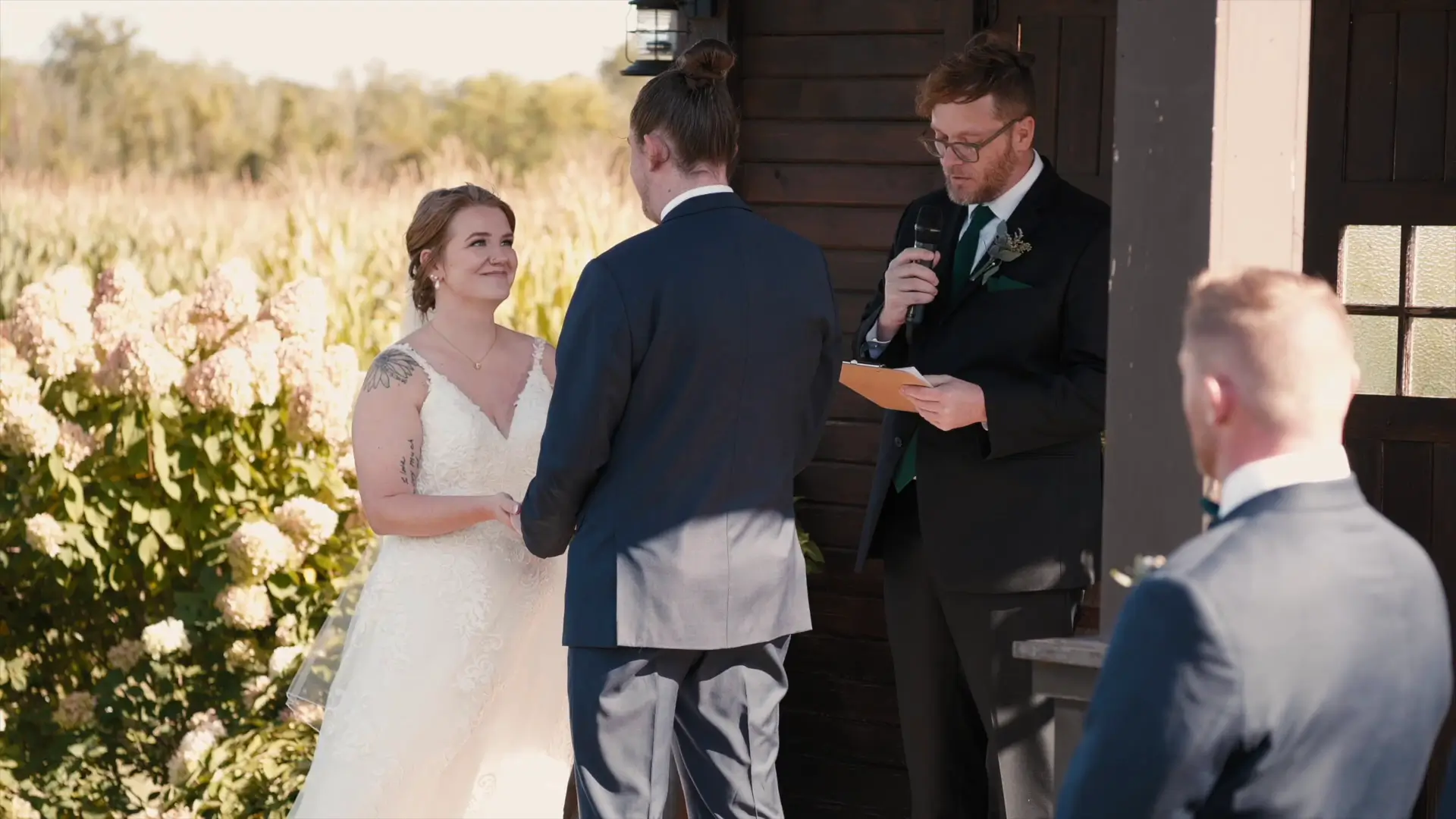 Bride and groom hold hands during an outdoor wedding ceremony at elegant Barn 1888. An officiant reads from notes while a groomsman watches in the foreground. Lush greenery and flowers surround them.
