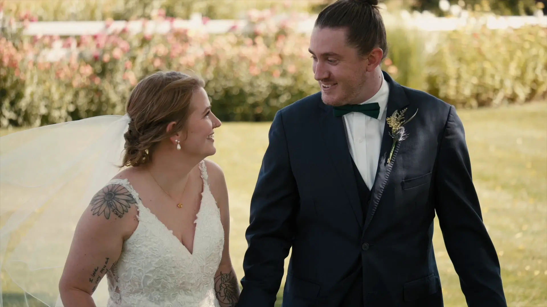 Bride Taylor and groom Josh smile at each other while walking outdoors in a garden setting at Barn 1888 in Hopkins, Michigan. She is wearing a lace dress, and he is in a suit with a dark green bow tie. Flowers are visible in the background.
