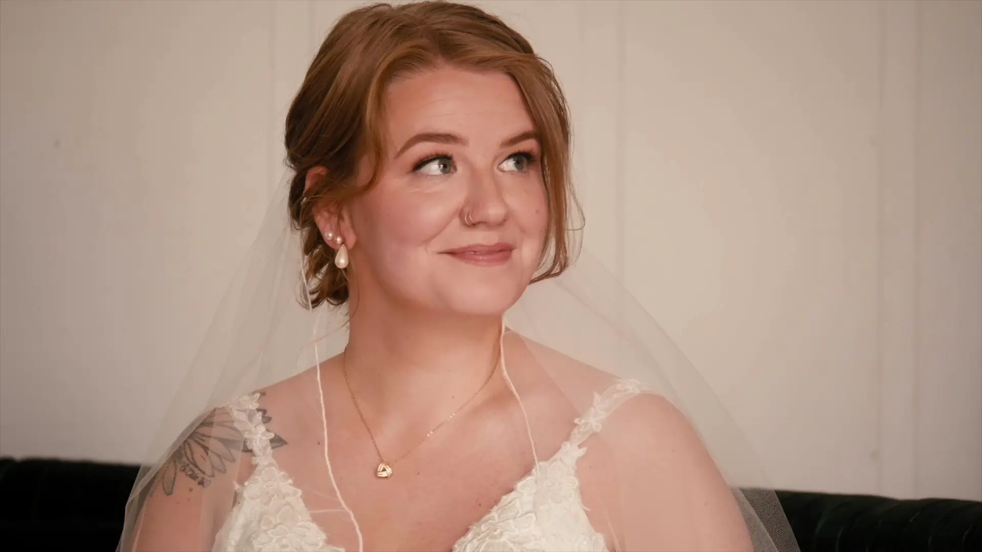 Bride Taylor smiles while wearing a lace wedding dress and veil, sitting in the bridal suite at Barn 1888 in Hopkins, Michigan. She's adorned with pearl earrings and a delicate necklace. Her expression is joyful and serene.