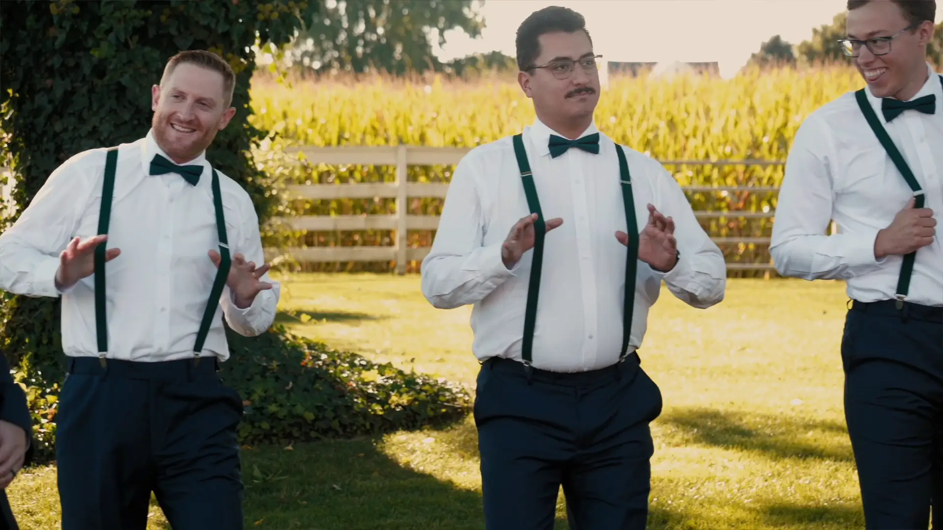 Three groomsmen wearing white shirts, bow ties, and suspenders enjoy a lighthearted moment outdoors at Barn1888 Events in Hopkins, Michigan. They are standing on grass with sunlight filtering through trees and a wooden fence in the background.