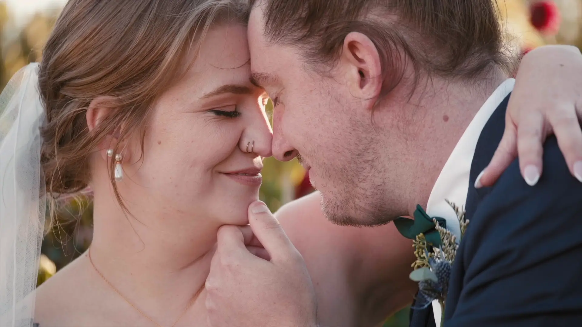 Bride and groom gently touching foreheads, smiling softly, with the bride wearing pearl earrings and a veil, outdoors with a warm ambiance.