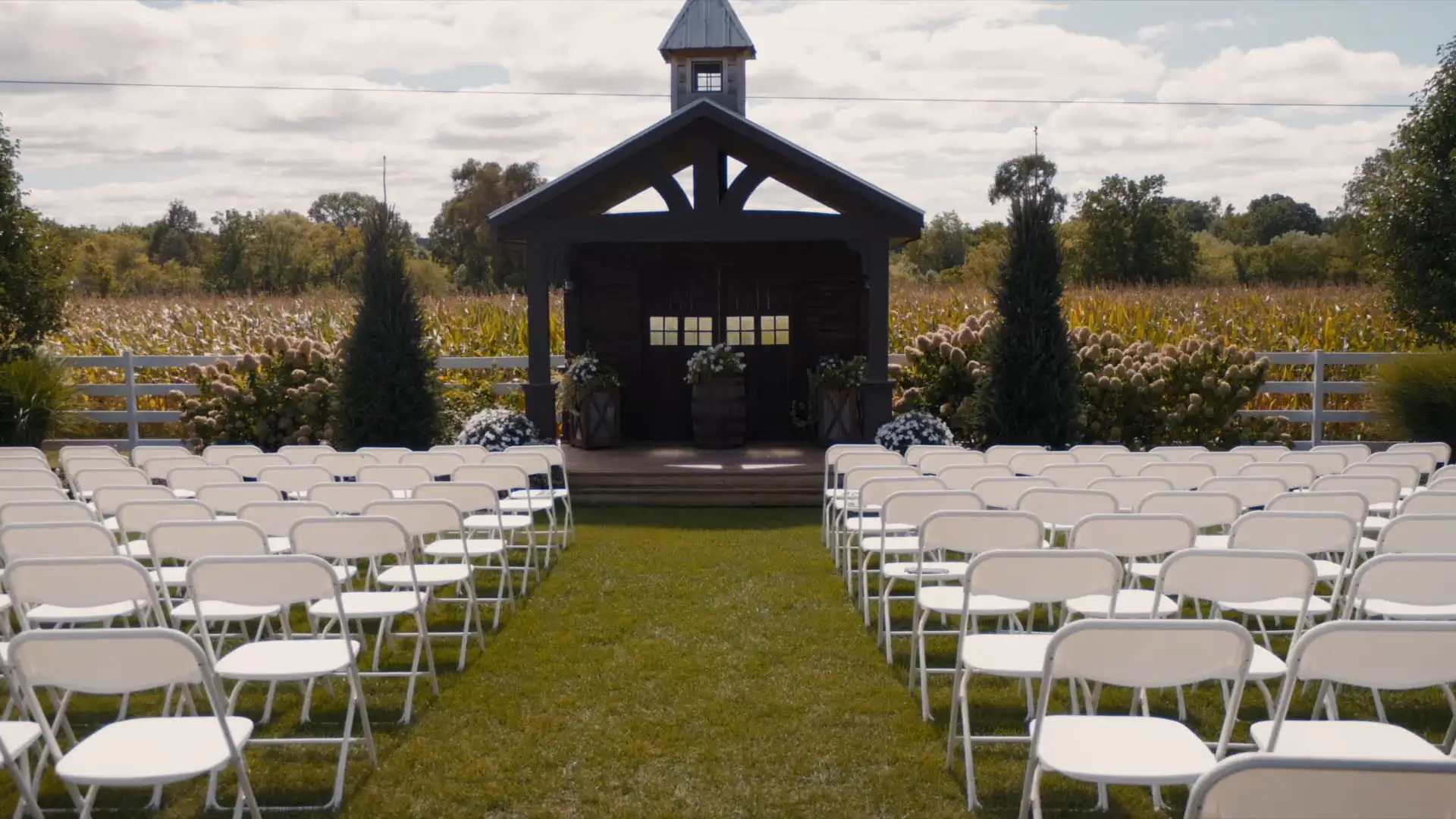 An elegant yet rustic wedding ceremony setup stands ready for the wedding at Barn1888 Events in Hopkins, Michigan. Rows of white folding chairs lead to a barn-like wooden pavilion, surrounded by greenery and fields, under a partly cloudy sky.