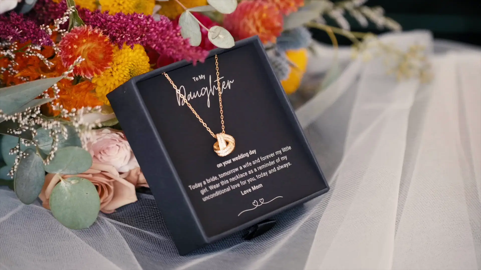 A delicate necklace in a black box with an engraved message "To My Daughter on your wedding day" is displayed against a background of vibrant flowers and soft white fabric.