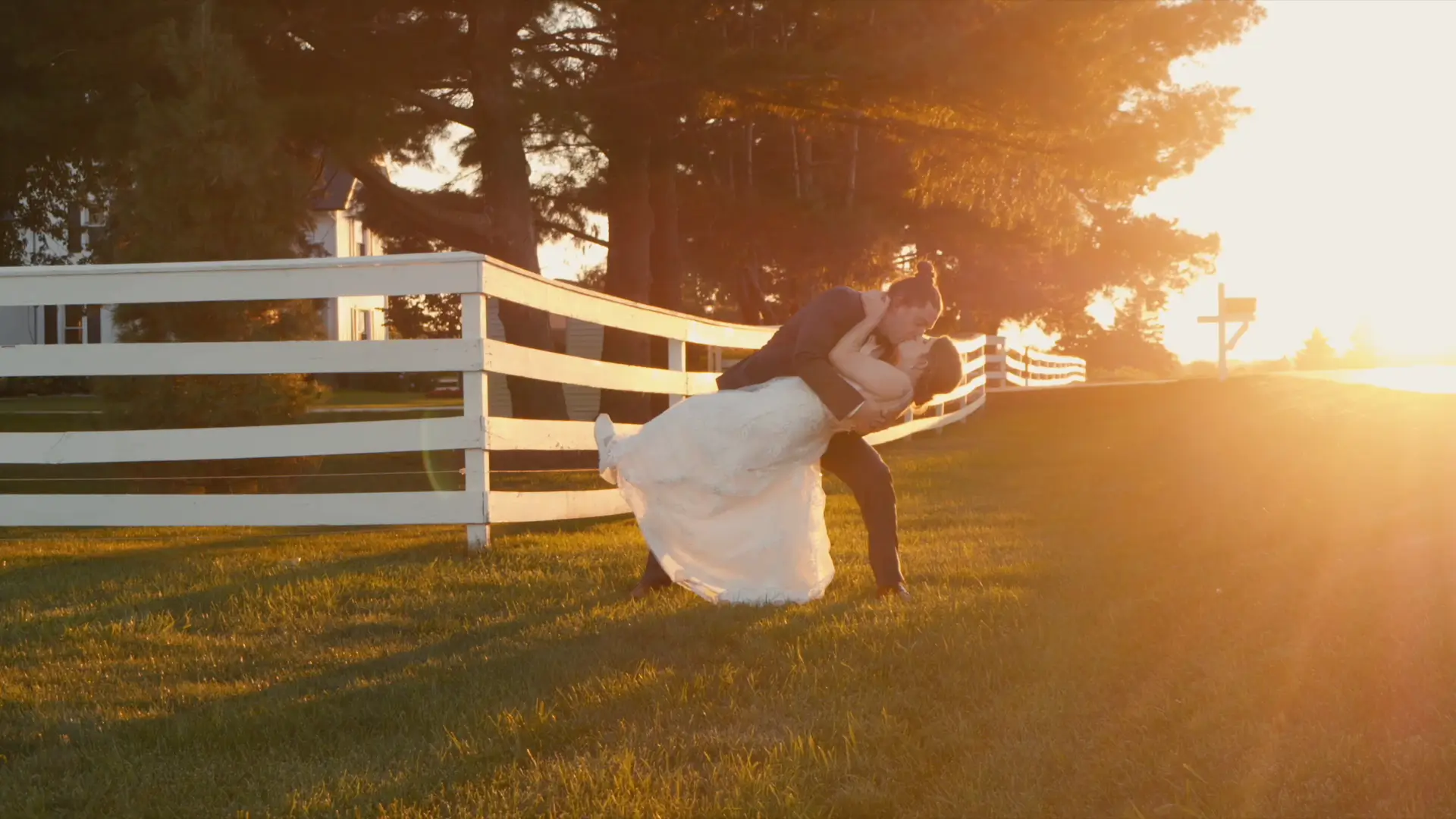 Bride and groom share a romantic dip kiss during a warm Michigan golden hour, with a white fence and tall trees in the background. The warm glow of the setting sun casts a romantic hue over the scene, captured cinematically for their wedding film.