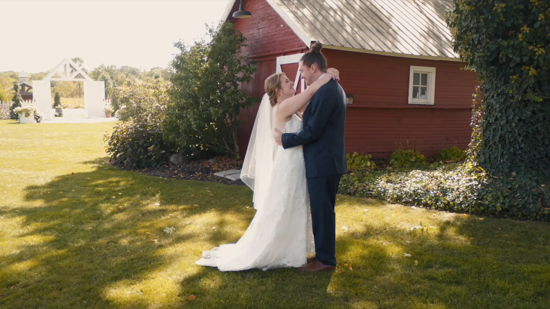 Bride and groom embrace outdoors in front of a red barn at Barn 1888 Events in Hopkins, Michigan. She wears a white gown and veil, and he wears a navy suit. The scene is sunny with greenery around.