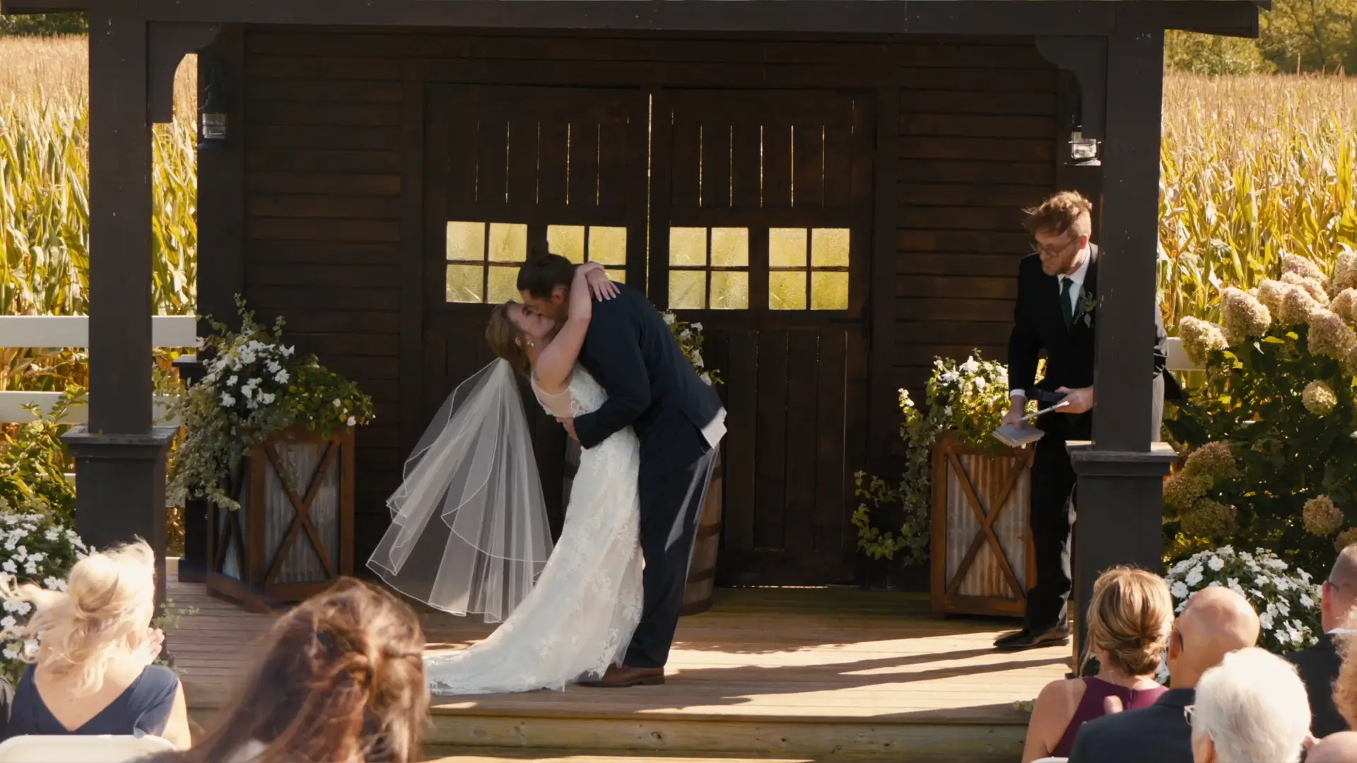 In this wedding videography moment at Barn 1888, Michigan, the newlyweds share their first kiss as a married couple on a rustic outdoor platform, surrounded by lush greenery and applauding guests.