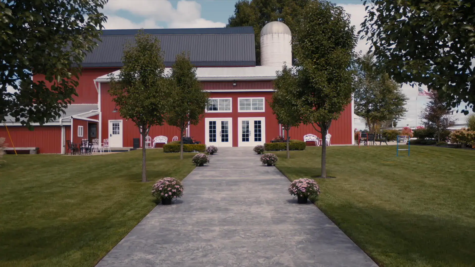 A wide walkway lined with trees and potted flowers leads to a large red barn with white-framed windows and doors at Barn1888 Events in Hopkins, Michigan. This still sets the scene for an elegant yet rustic wedding, as captured by a wedding videographer.