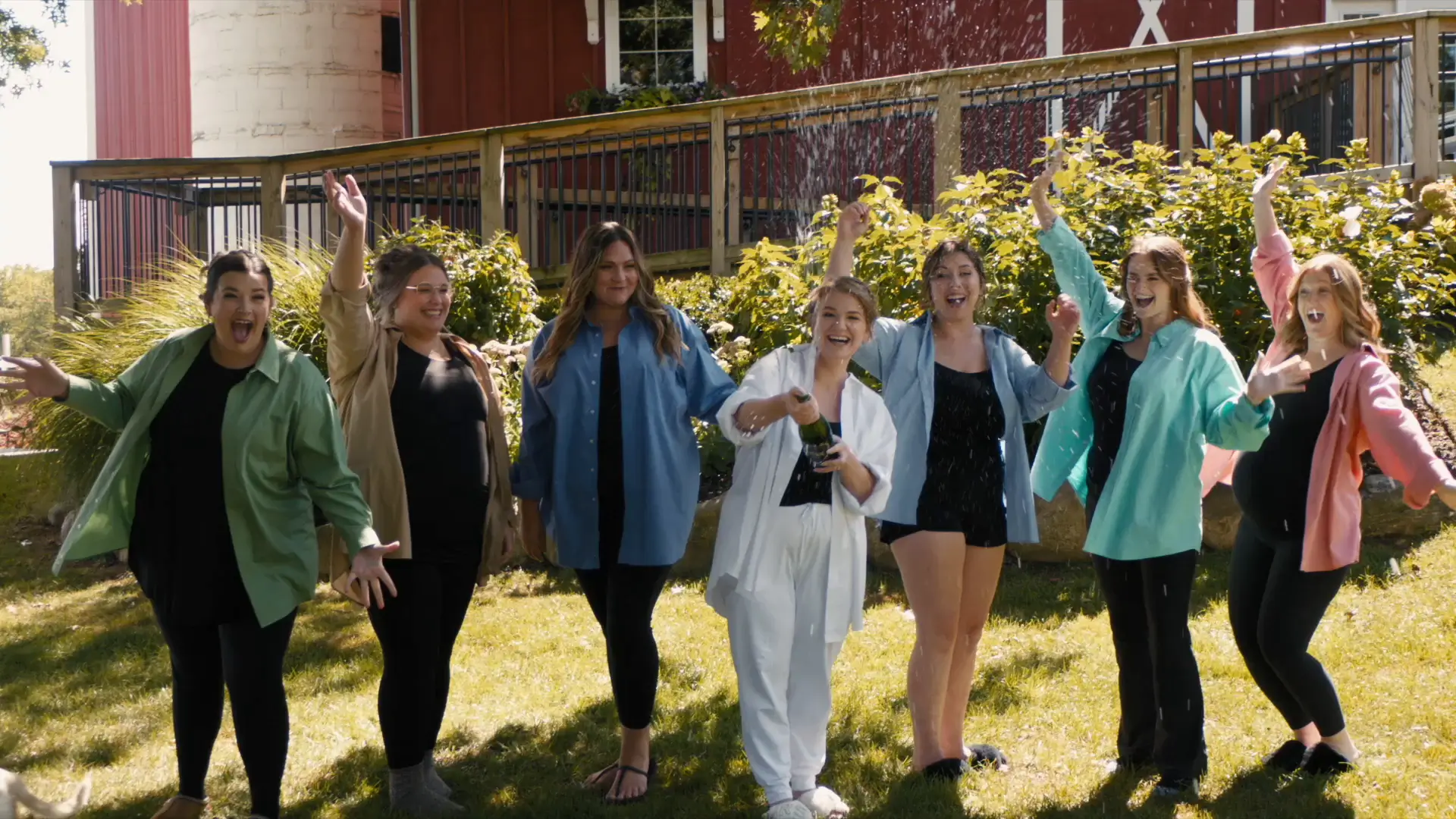 A group of bridesmaids cheer as the bride in white pops champagne outdoors at Barn 1888 Events. They wear coordinating casual, colorful attire under sunny skies, with a red barn and greenery in the background.