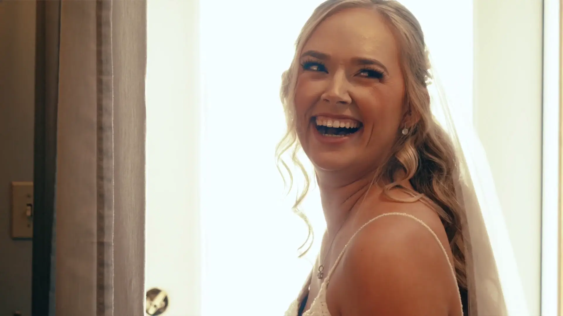 Bride Caroline smiles broadly in a spaghetti-strap dress, soft light catching her wavy hair and a veil over her shoulder as she looks back near a doorway.