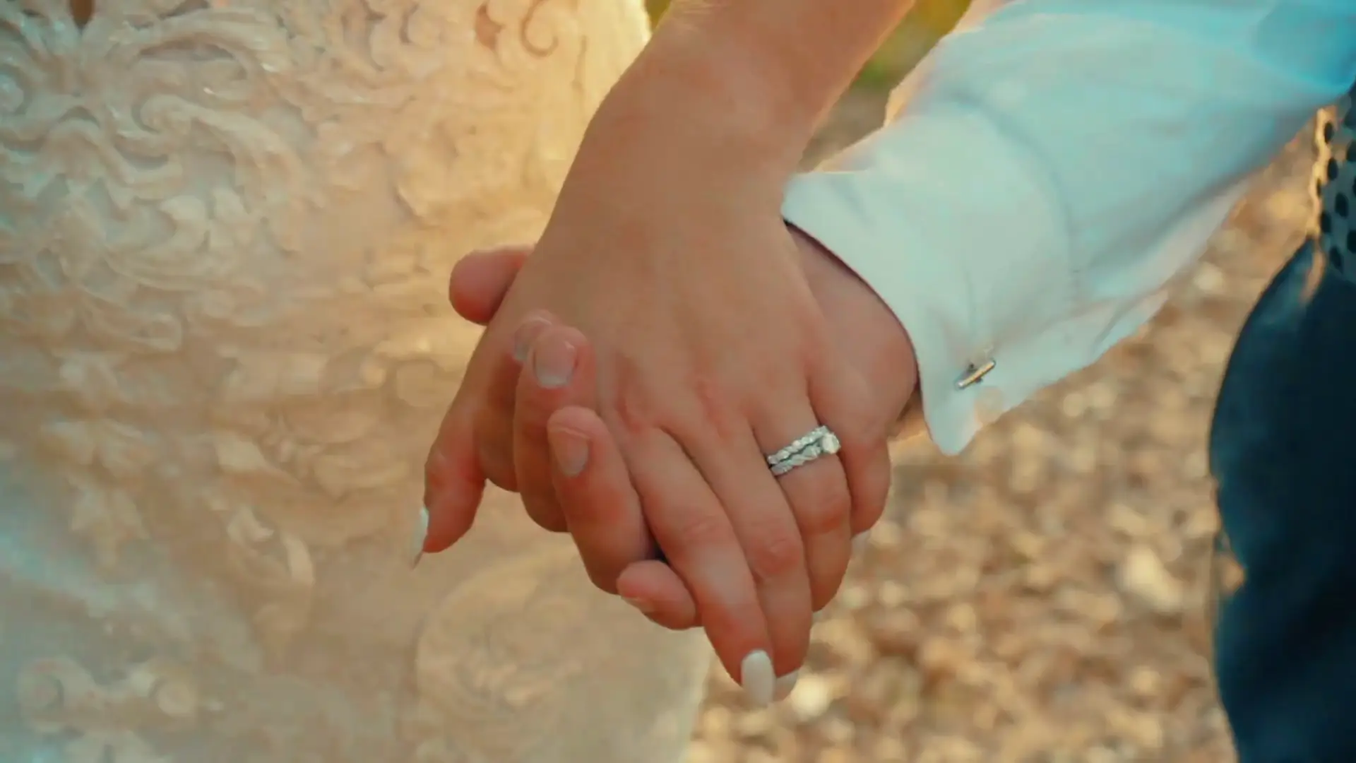 In this wedding videography still from Caroline and Matt's Michigan celebration, the bride and groom hold hands, highlighting the bride's intricate lace dress and sparkling ring. The sunlit scene captures their connection and joy.