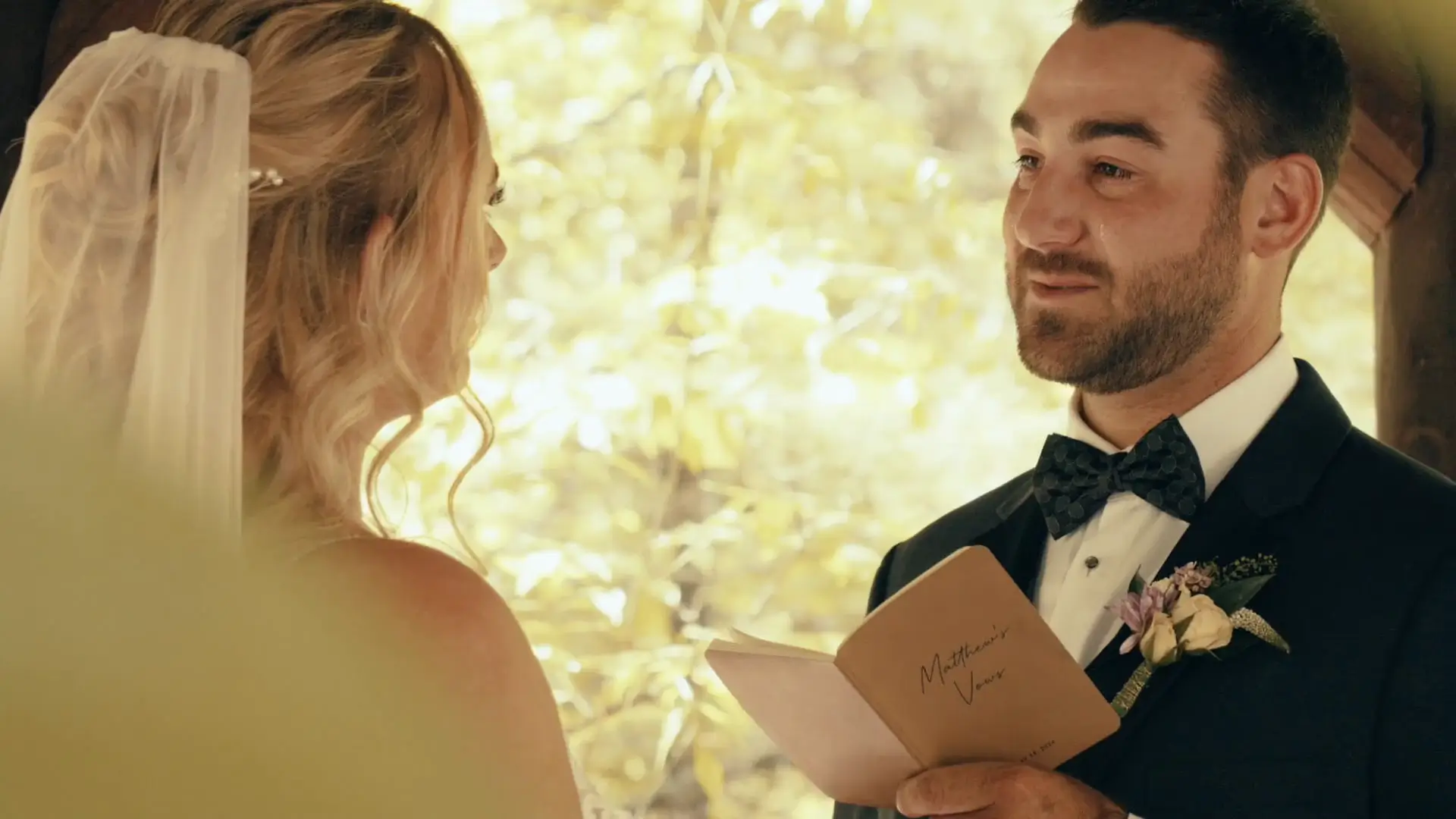 An emotional groom reads personal vows to his bride during their wedding ceremony at BlueBridge Events Centre. The bride, seen from behind with a veil, is listening intently amid a warm, glowing backdrop of soft, yellow Michigan foliage.