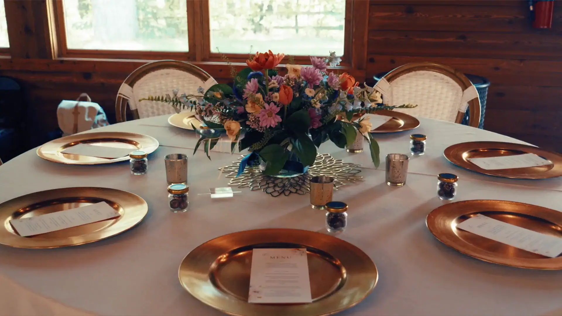 A round table set for a wedding at Blue Bridge Events Centre in Traverse City, Michigan. It features gold plates, menus, small jars, and a vibrant floral centerpiece with orange and pink flowers, creating a festive and elegant atmosphere.
