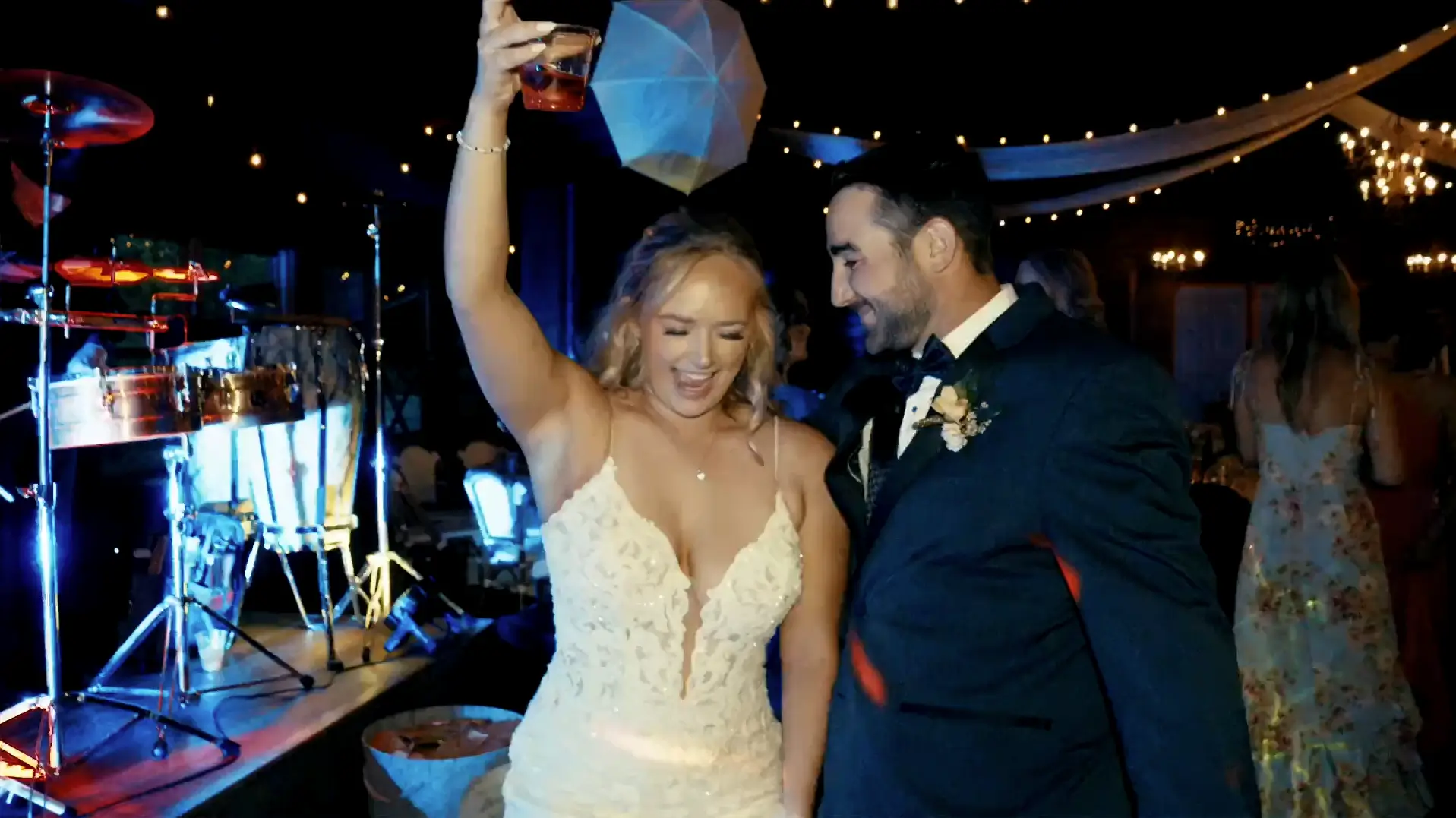 A bride in a detailed lace dress joyfully raises a drink while dancing with the groom in a suit, surrounded by party lights and musical instruments in a lively wedding reception atmosphere.