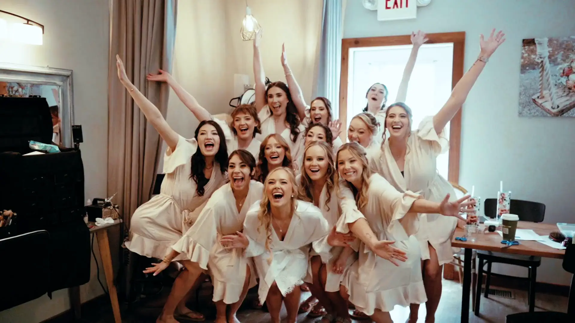 Group of bridesmaids in matching robes pose with arms raised, smiling in a bright room. This bridal party prep scene, full of excitement and celebration, is featured before Caroline and Matt's ceremony in their elegant wedding video.