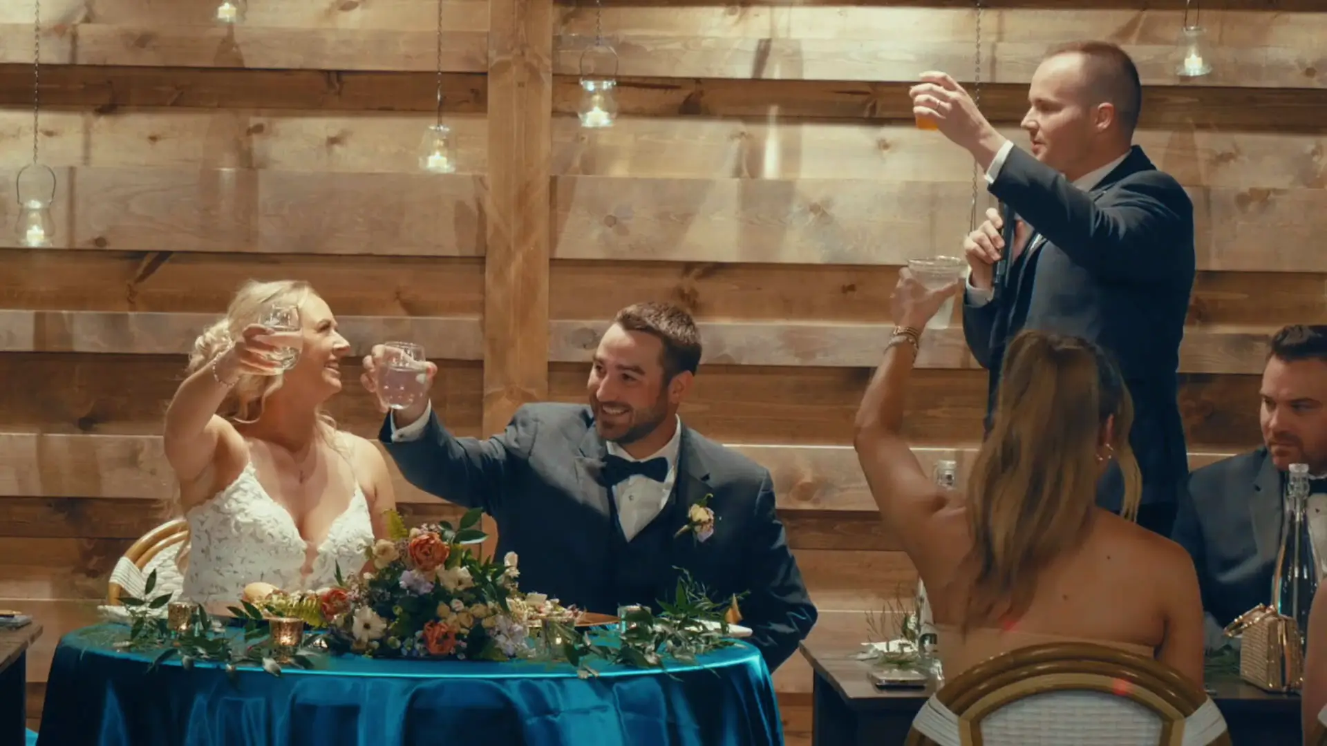 Caroline and Matthew raise glasses in a joyful toast, surrounded by loved ones under rustic wood decor. A perfect capture from the Michigan celebration by the videographer.