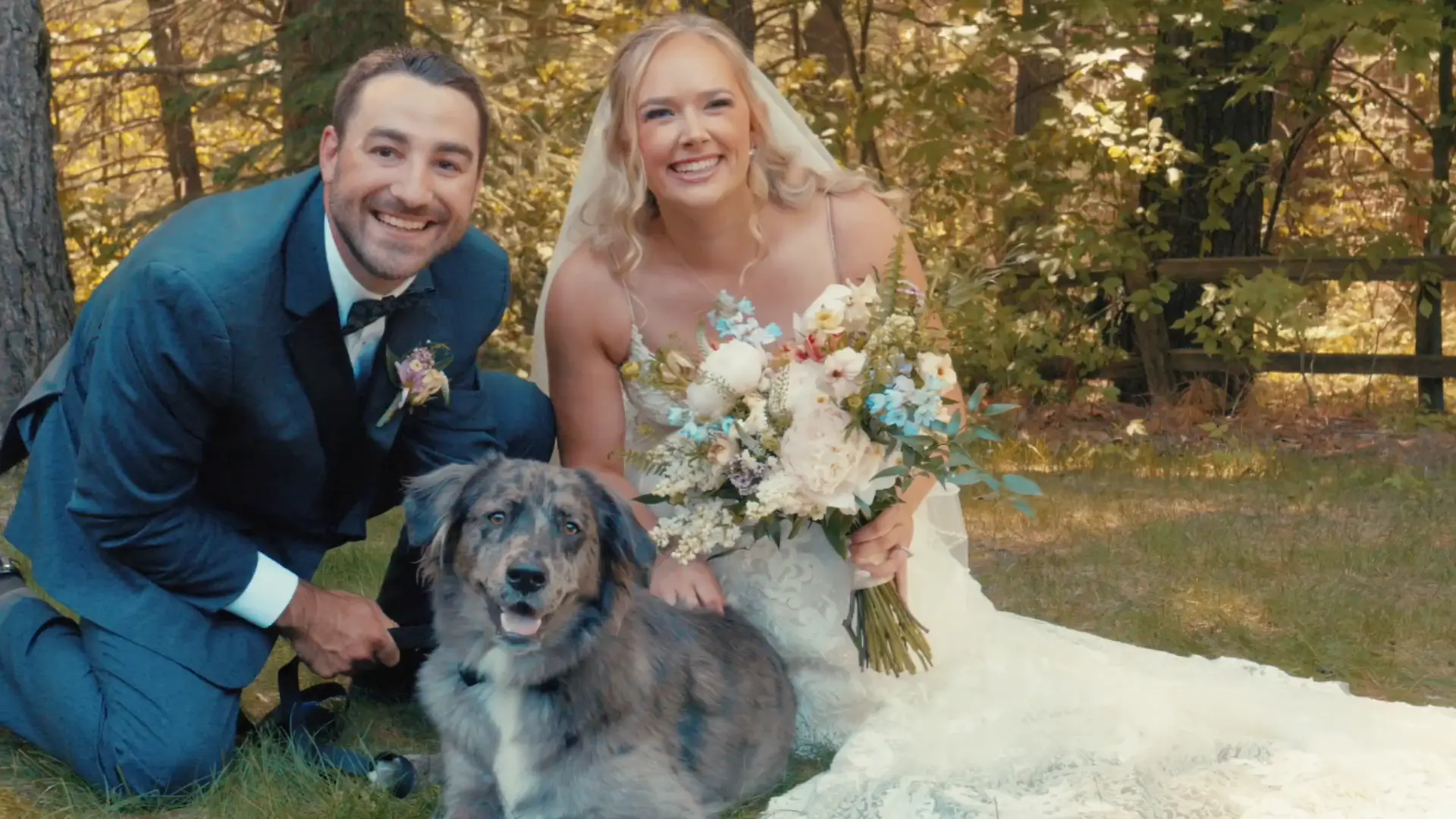 Bride and groom smiling with their dog, kneeling on grass at BlueBridge Events in Grawn, Michigan. The bride holds a bouquet, and the scene is outdoors with trees in the background.