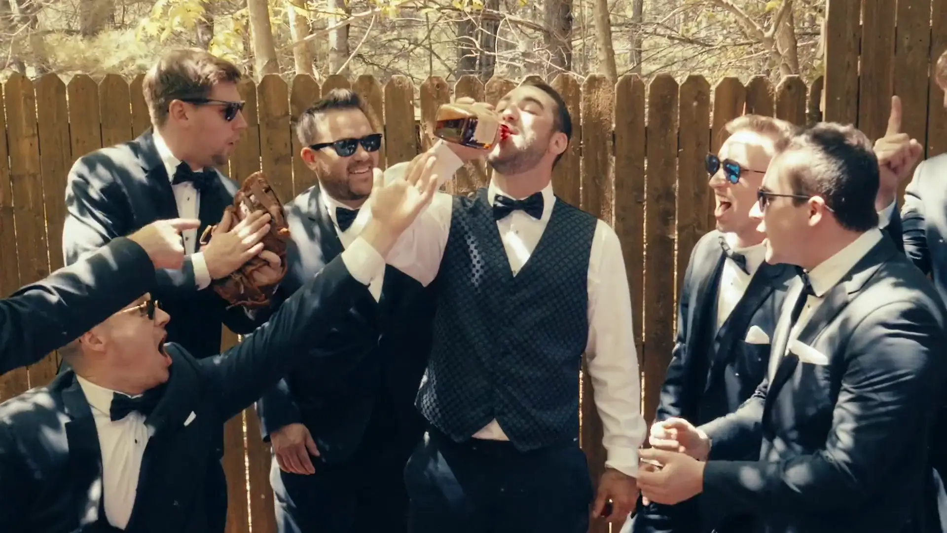 Men in tuxedos, some wearing sunglasses, gather outside by a wooden fence. The groom drinks from a bottle while others cheer him on, creating a lively, celebratory scene captured by their wedding videographer.