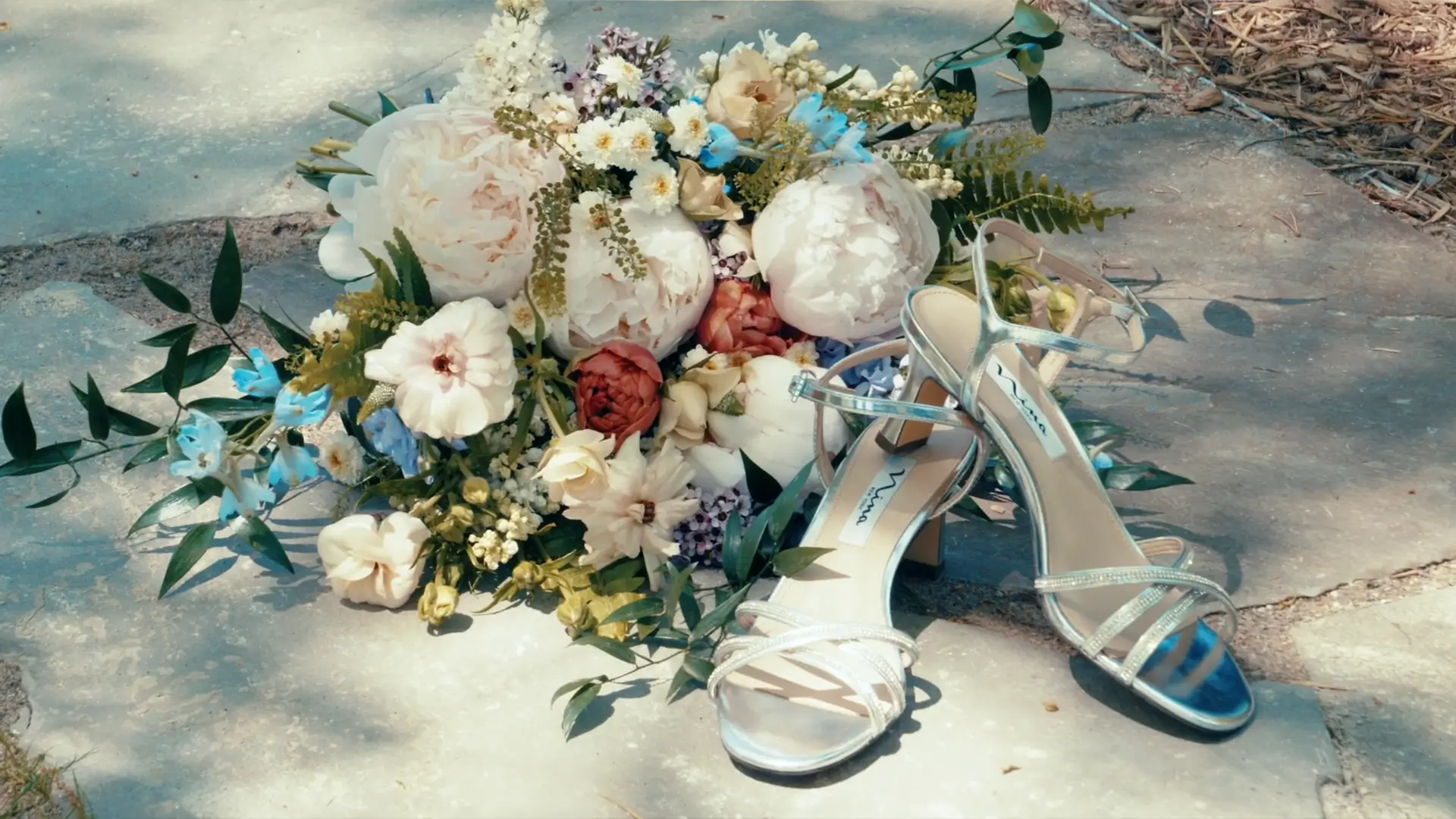 A delicate bridal bouquet with white, pink, and blue flowers lies on a stone pavement alongside elegant silver strappy high-heeled sandals. Sunlight and shadows create a warm, inviting atmosphere.