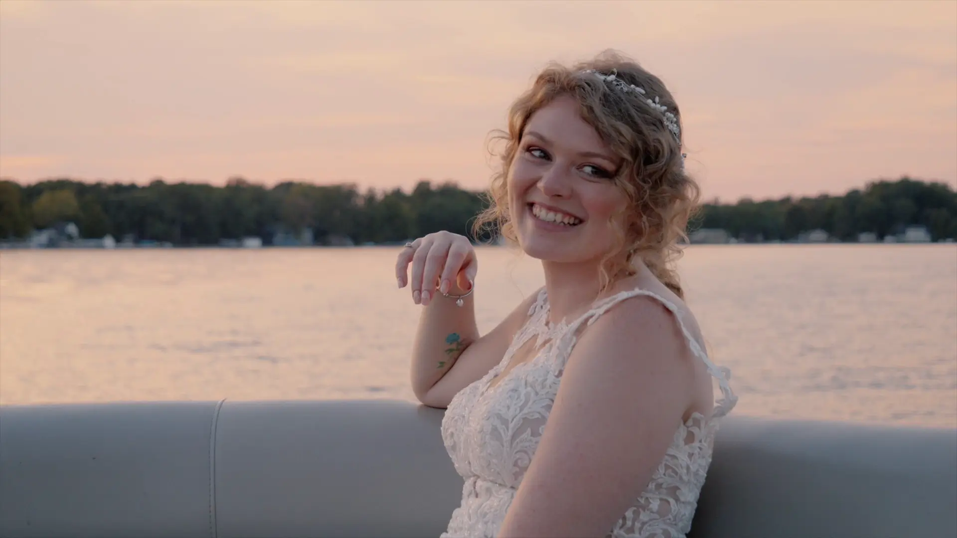 A bride in a lace dress smiles joyfully on a boat during sunset, with a beautiful lake and tree-lined shore in the background. Captured by their wedding videographer.