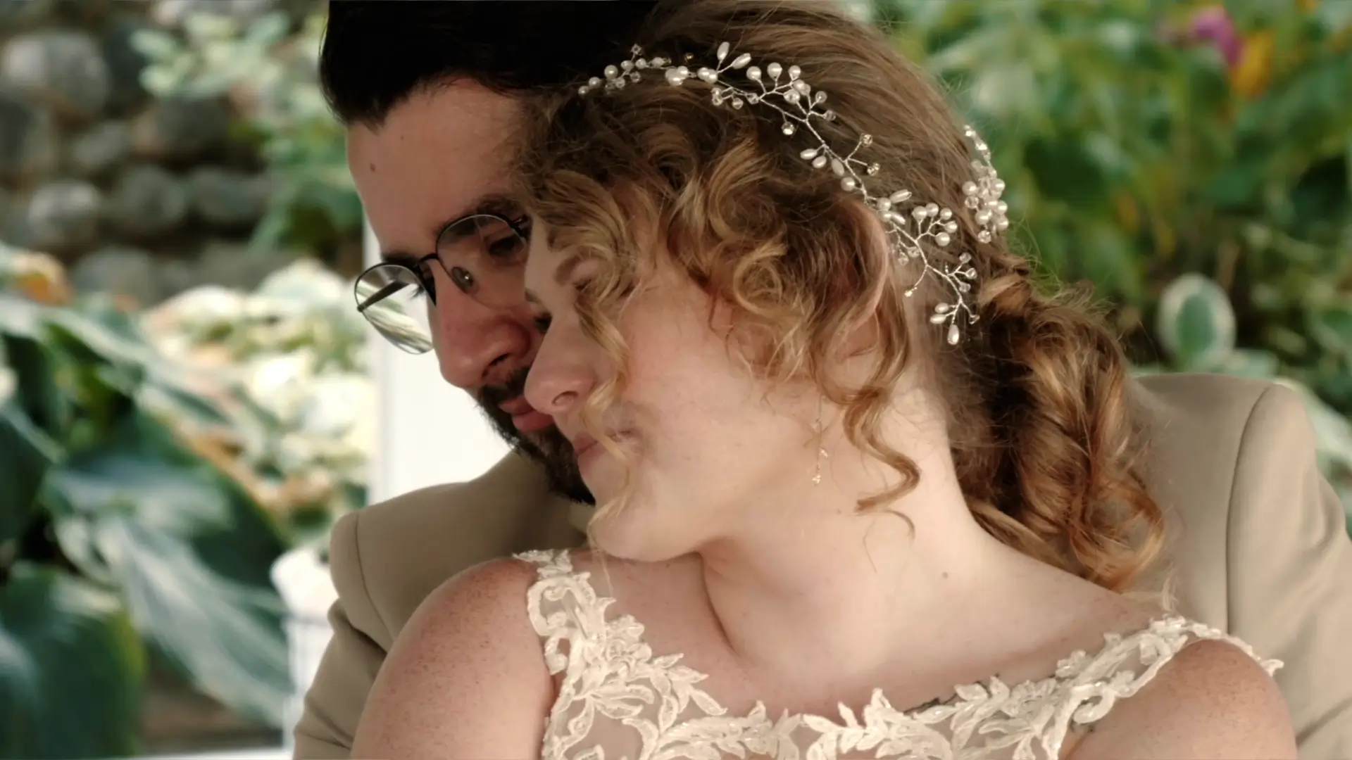 In this still from their wedding video, a bride with curly hair and a pearl headpiece leans affectionately against her partner, captured by their wedding videographer.