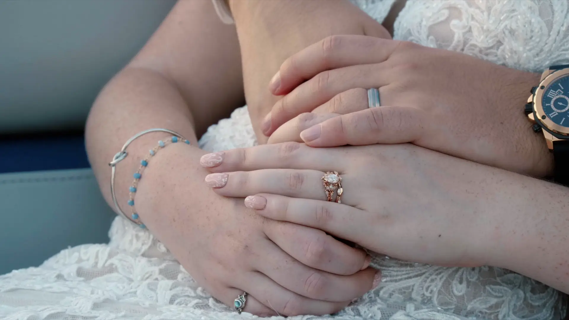 Close-up of hands with wedding rings resting on lace fabric, featuring a bejeweled bracelet and a watch.