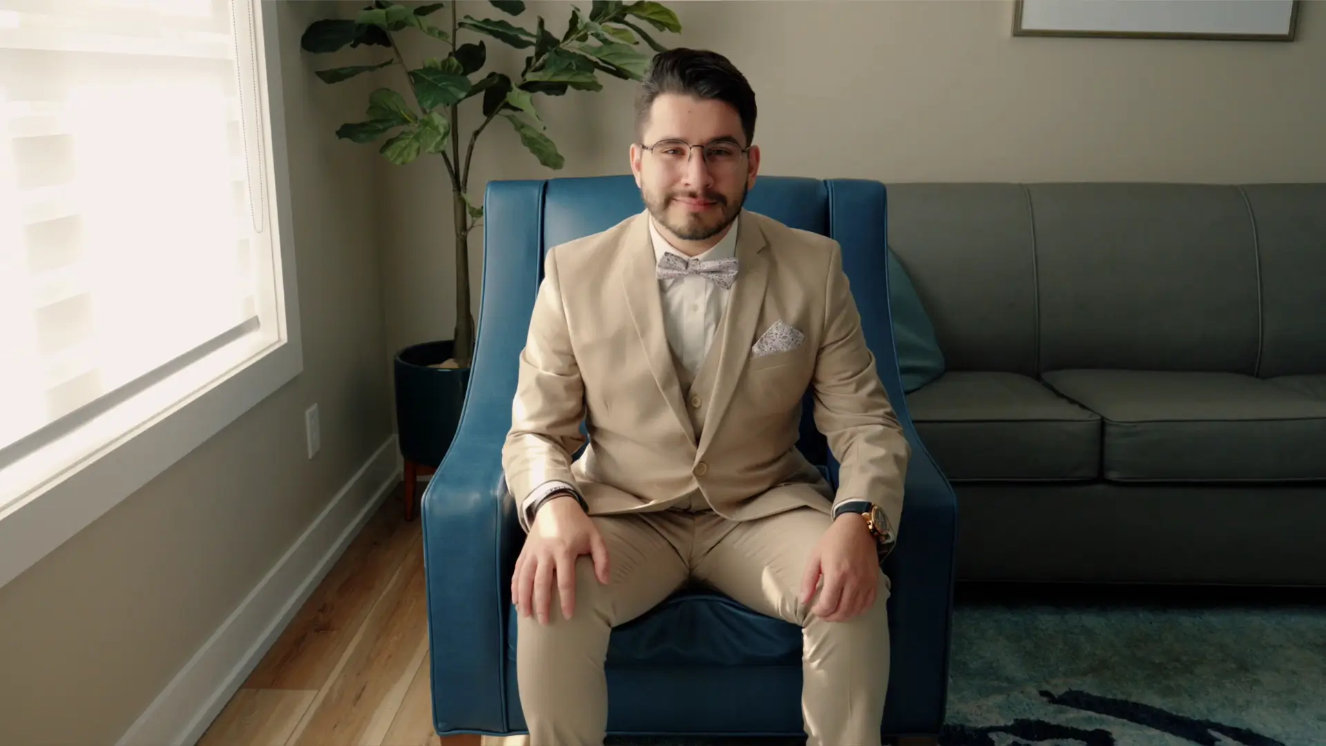 A man in a beige suit with a bow tie sits on a blue chair in a well-lit room.