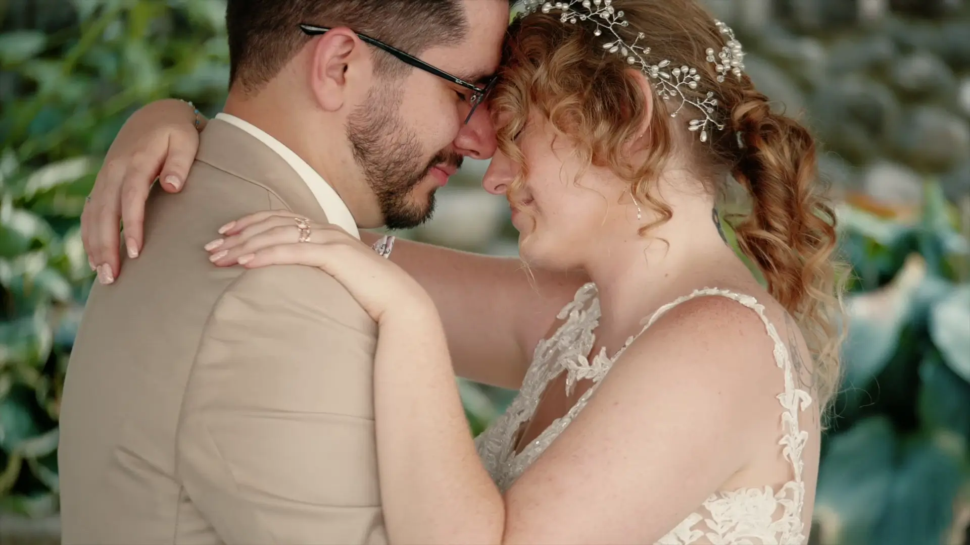 A bride and groom share an intimate moment, touching foreheads and holding each other closely. She wears a delicate hairpiece and lace dress, while he is in a beige suit. Captured by their wedding videographer amidst green foliage.
