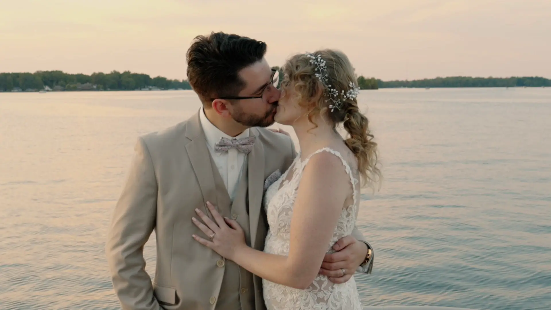 In this still from their wedding video, a couple shares a romantic kiss by a tranquil waterfront during sunset, the bride in a lace gown and the groom in a tan suit.