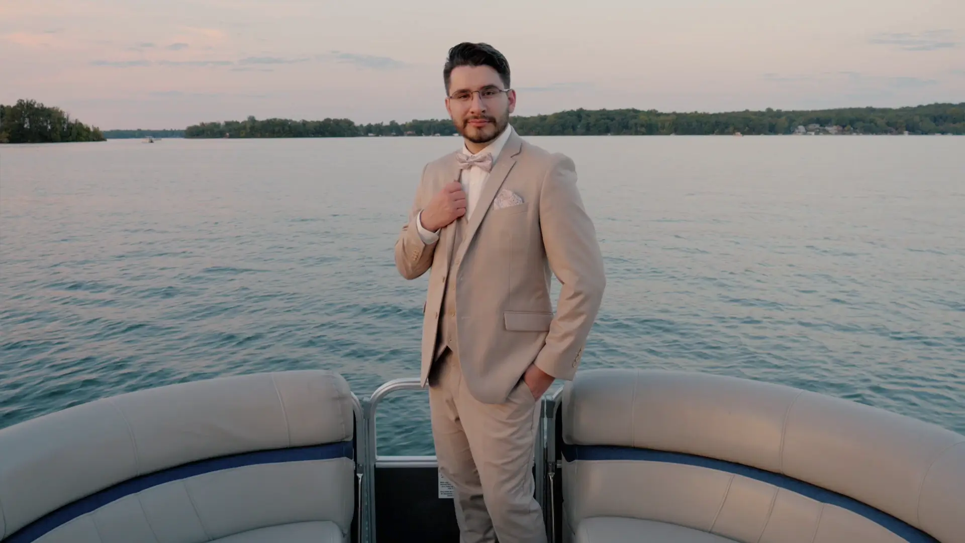 A groom in a beige suit poses confidently on a boat with a scenic lake and tree-lined horizon in the background during sunset, captured by their wedding videographer.