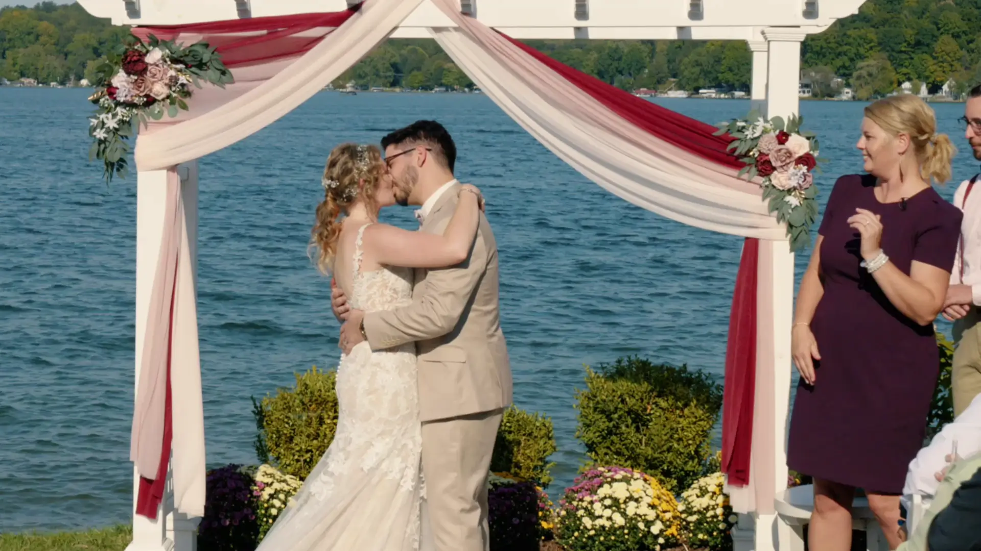 In this still from their wedding video, a bride and groom share a kiss under a floral arch by a lakeside, captured by their wedding videographer.