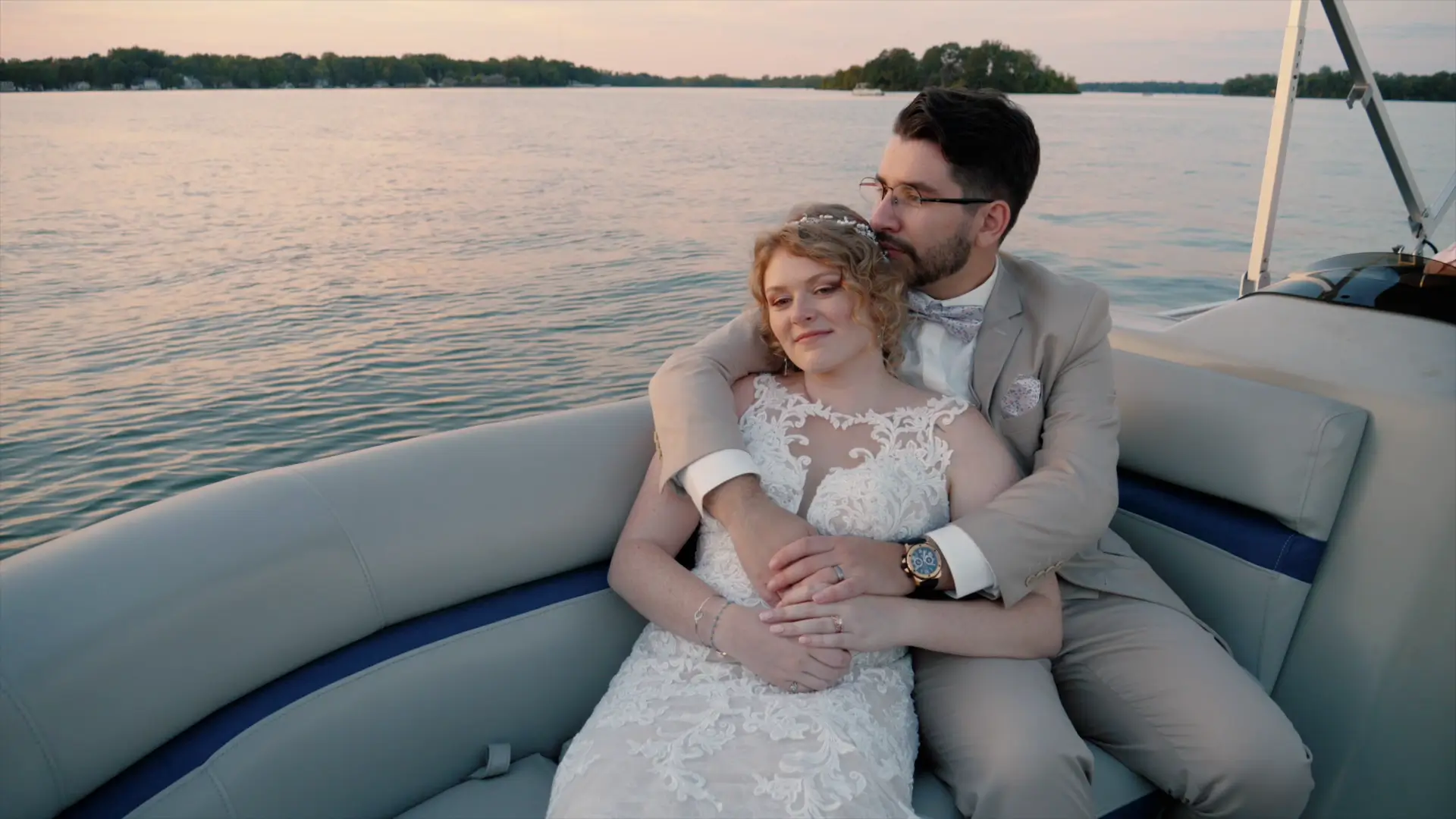 A bride and groom embrace on a boat at sunset, with water and trees in the background. The groom is dressed in a suit and the bride in a lace gown, creating a serene and intimate moment captured by their wedding videographer.