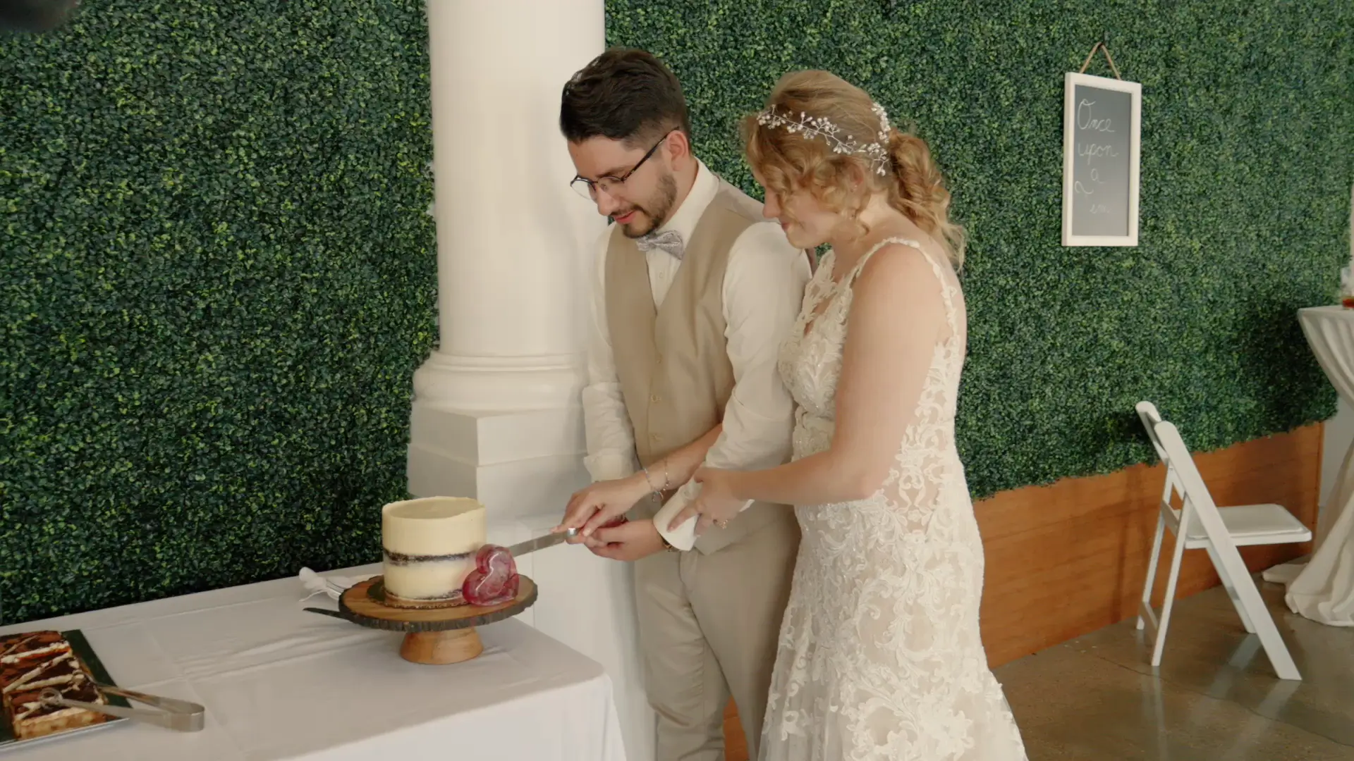 A bride and groom in formal attire are cutting a small wedding cake together, with a green foliage wall in the background.