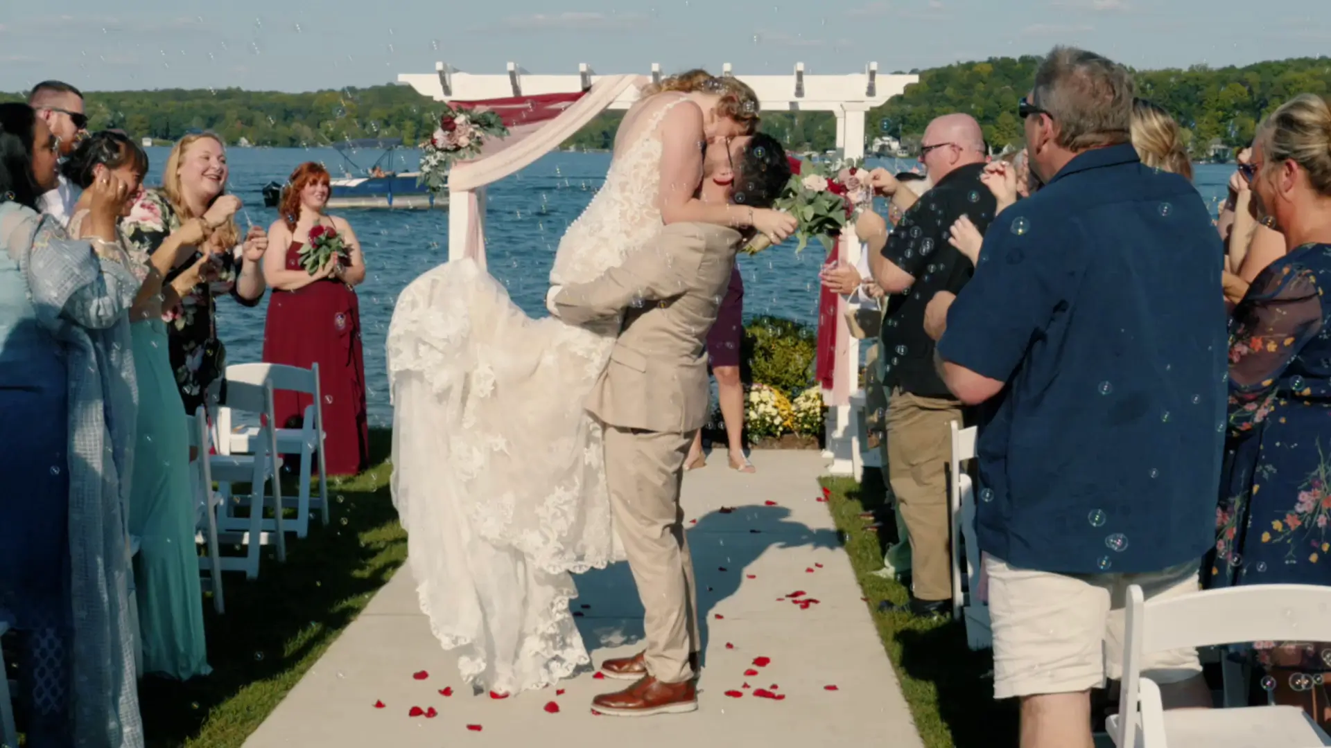A groom lifts the bride for a kiss surrounded by guests blowing bubbles during an outdoor wedding ceremony by a lakeside, captured in their wedding video.