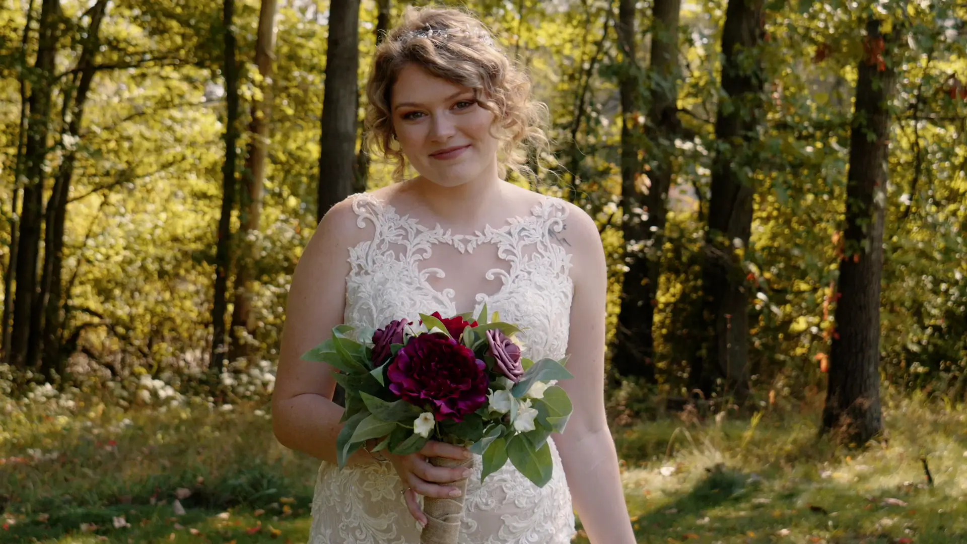 Woman in a white lace gown holding a bouquet of flowers, standing in a wooded area with sunlight filtering through the trees.