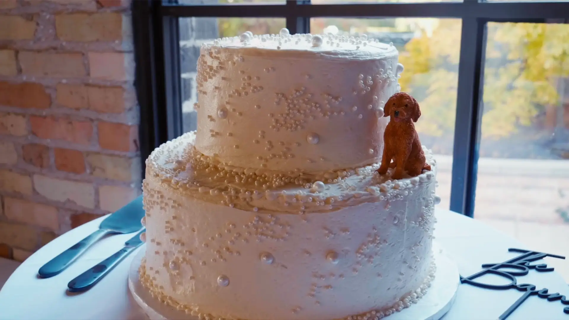 White wedding cake adorned with pearly decorations and a figurine of a brown dog on top, set against a backdrop of exposed brick and a window displaying vibrant Michigan foliage.