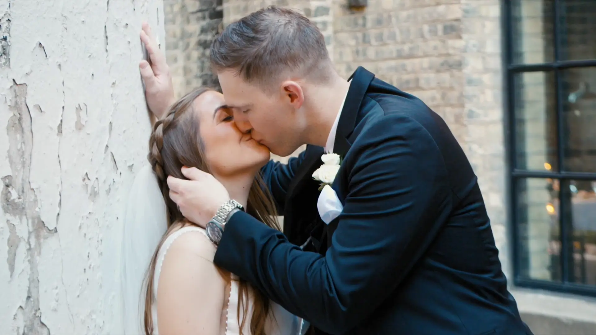 Bride and groom share a kiss against a textured wall. Groom gently holds bride's face. Bride has long braided hair. Both are in formal attire, with groom wearing a suit with a white rose boutonniere. Brick wall and window in the background.