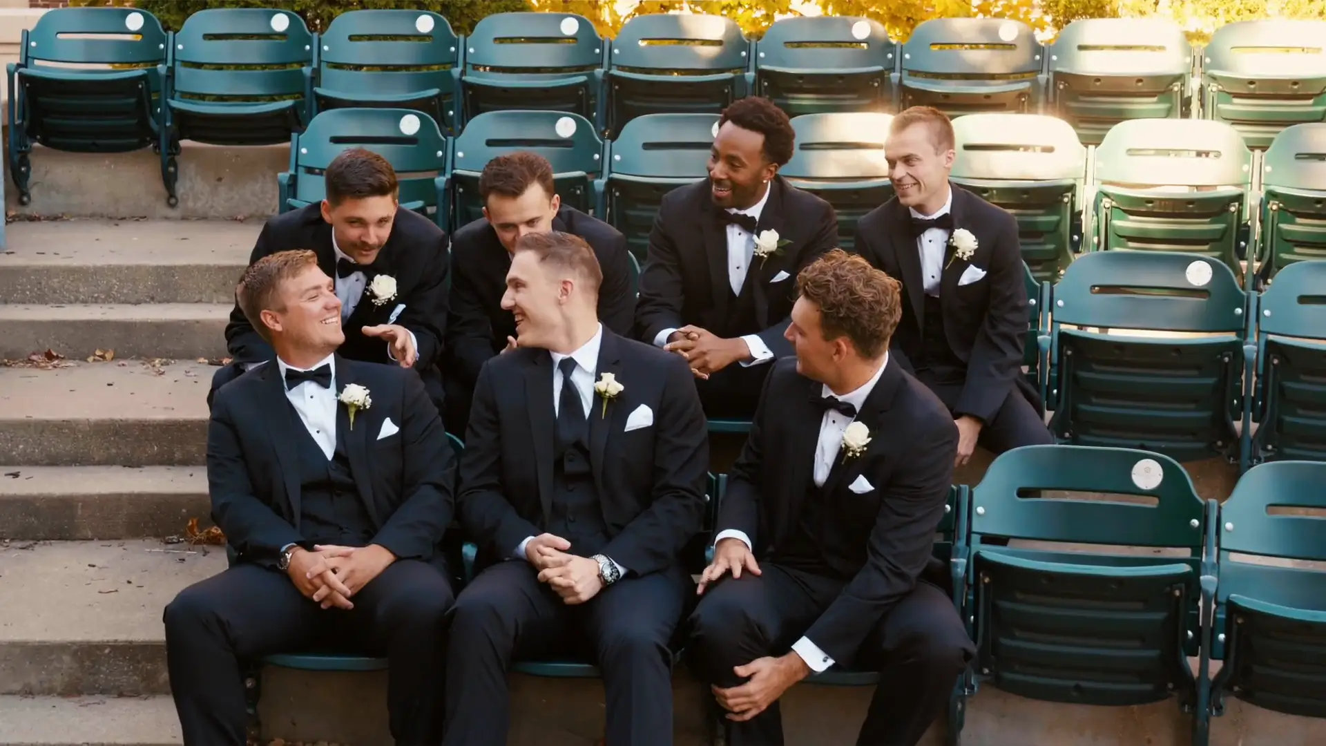 Groomsmen in tuxedos share laughter while seated on green stadium chairs at a Michigan venue, reflecting a joyful moment before Madison and Max's wedding in Grand Rapids, Michigan.