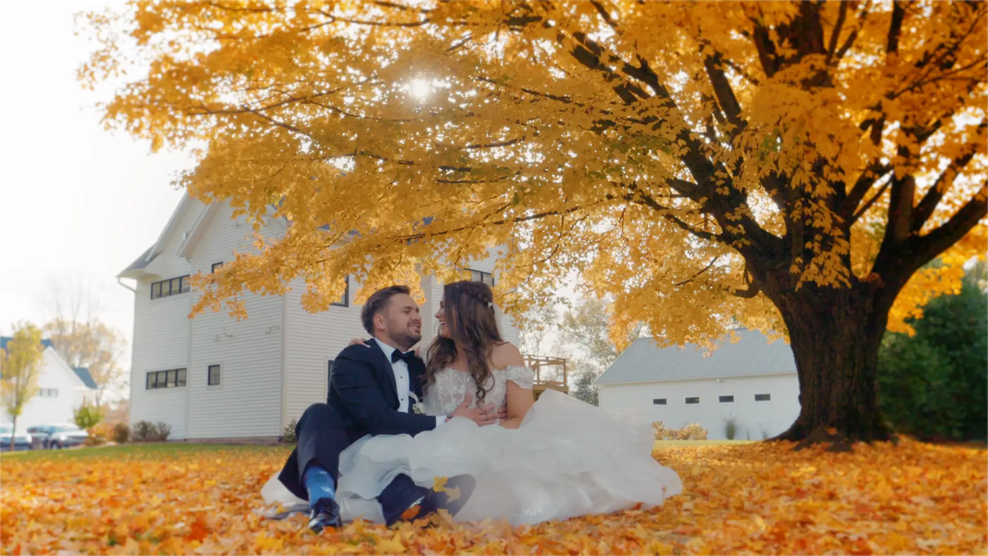 Bride and groom sit embraced under a large tree with vibrant yellow leaves, smiling at each other. A white building is visible in the background. Sunny day with autumn leaves covering the ground.