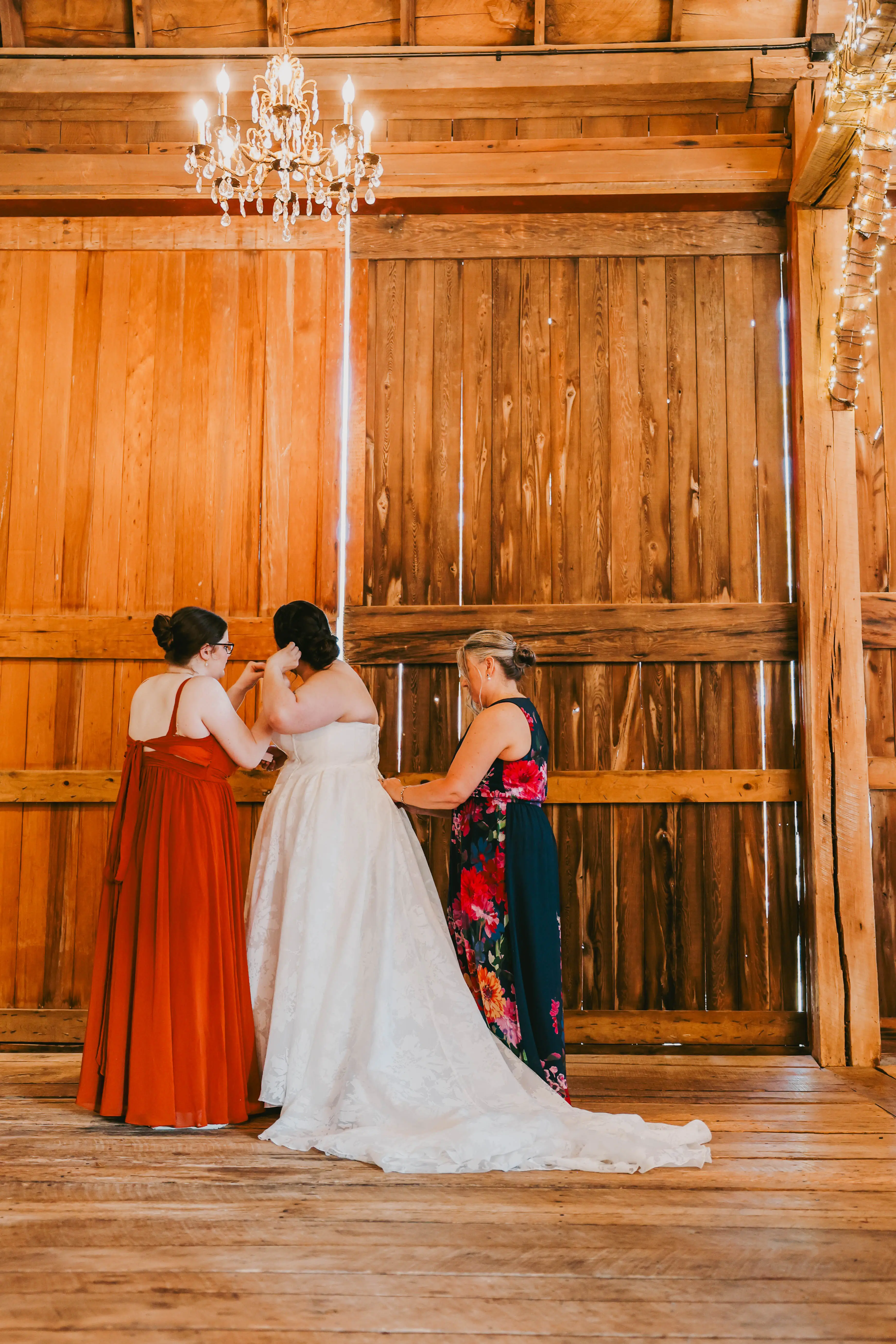 A bride in a white dress being assisted by two women in a wooden barn under a chandelier.