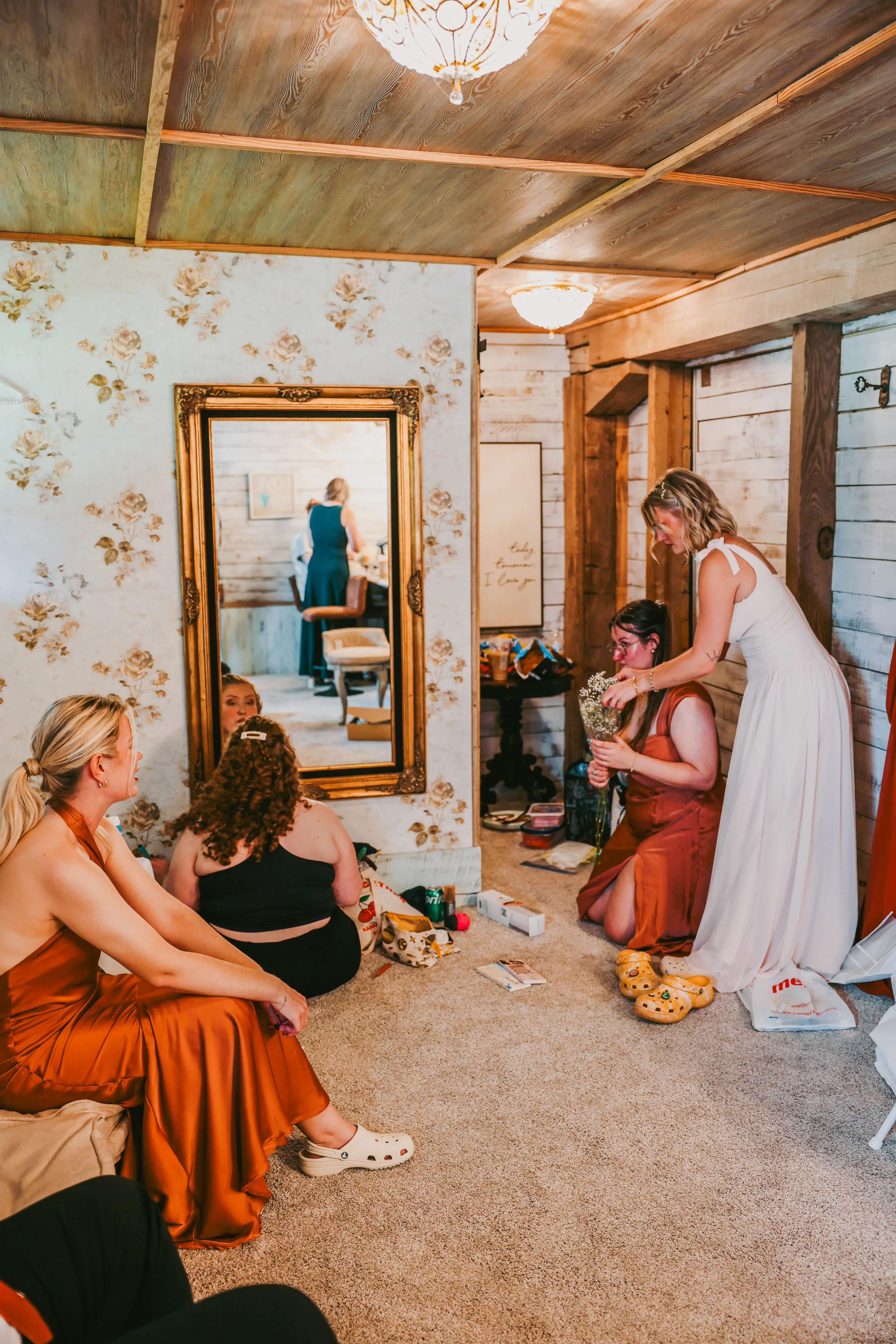A group of women in a cozy room getting ready for a wedding, with one adjusting another's dress, surrounded by various items and a large mirror reflecting the scene.