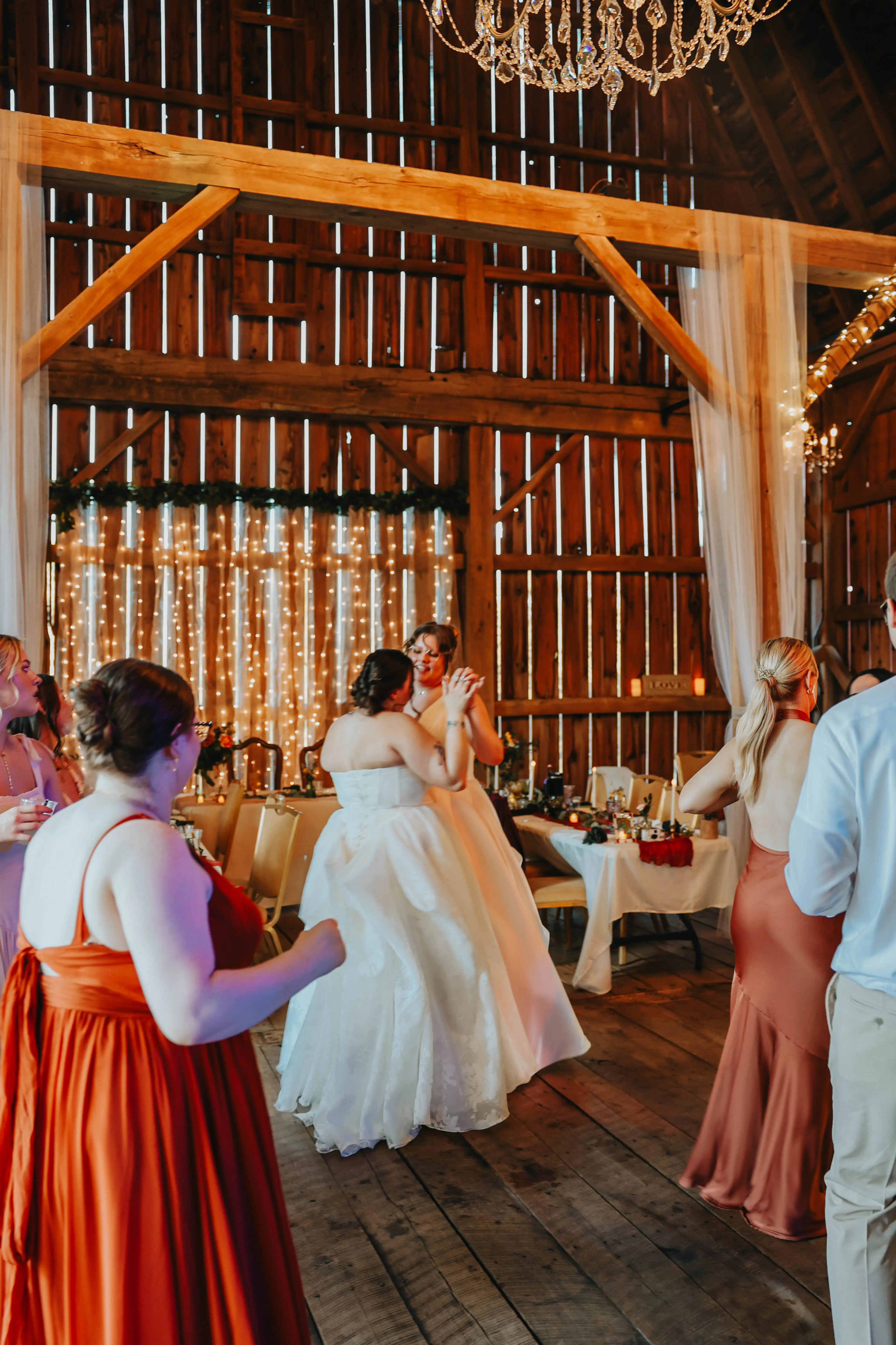 A couple in wedding attire sharing a dance at a wedding reception inside a rustic barn, surrounded by guests.