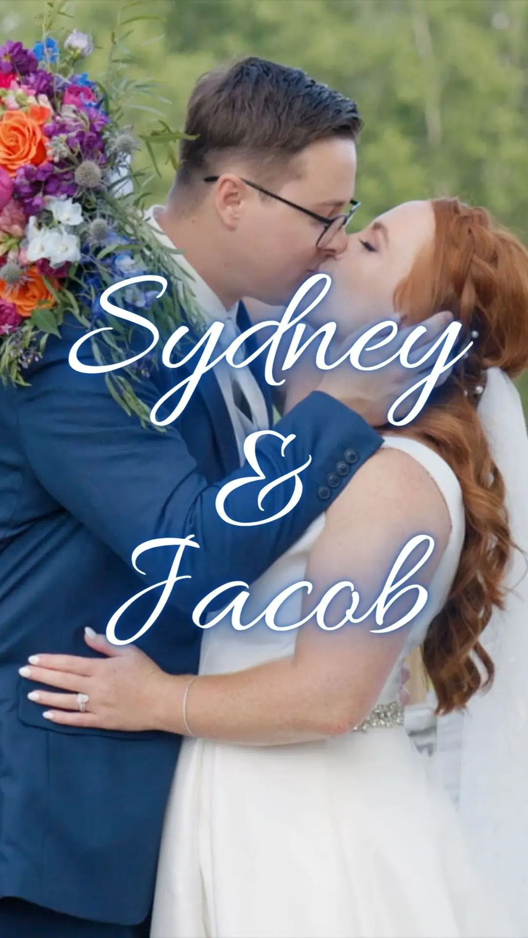 A couple kissing at their wedding, with the bride in a white dress holding a colorful bouquet and the groom in a navy suit. The background is a blurred green setting.