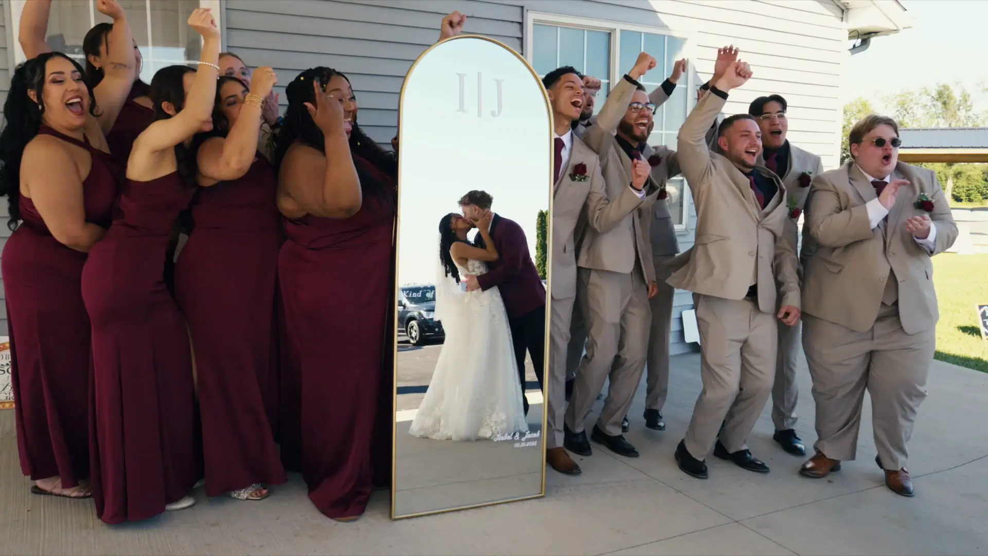 In this still from their wedding video, the bridal party cheers as the bride and groom kiss reflected in a large mirror.