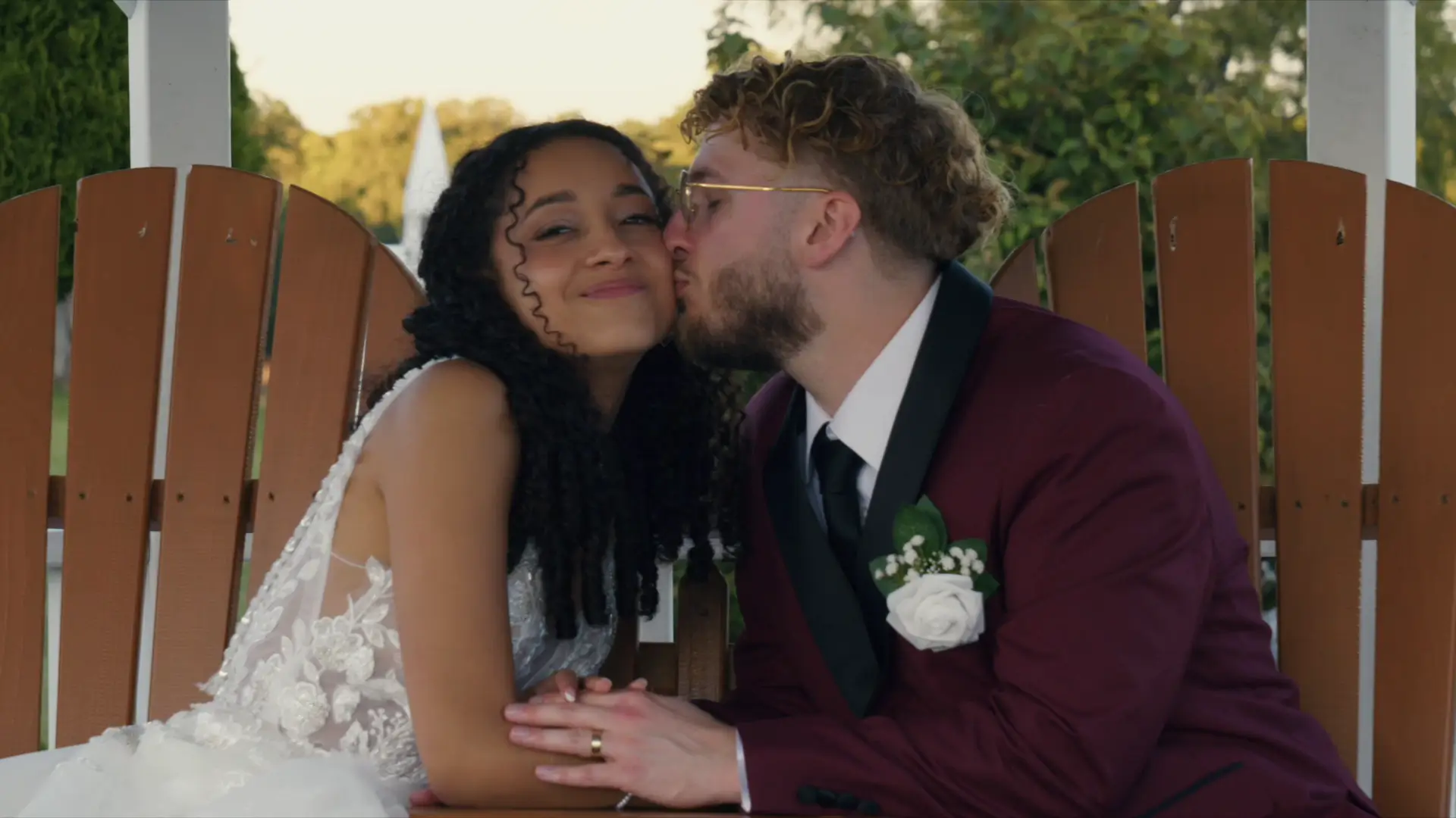 In this still from their wedding video, a groom in a burgundy suit leans to kiss his smiling bride in a lace gown, seated on wooden chairs outdoors.