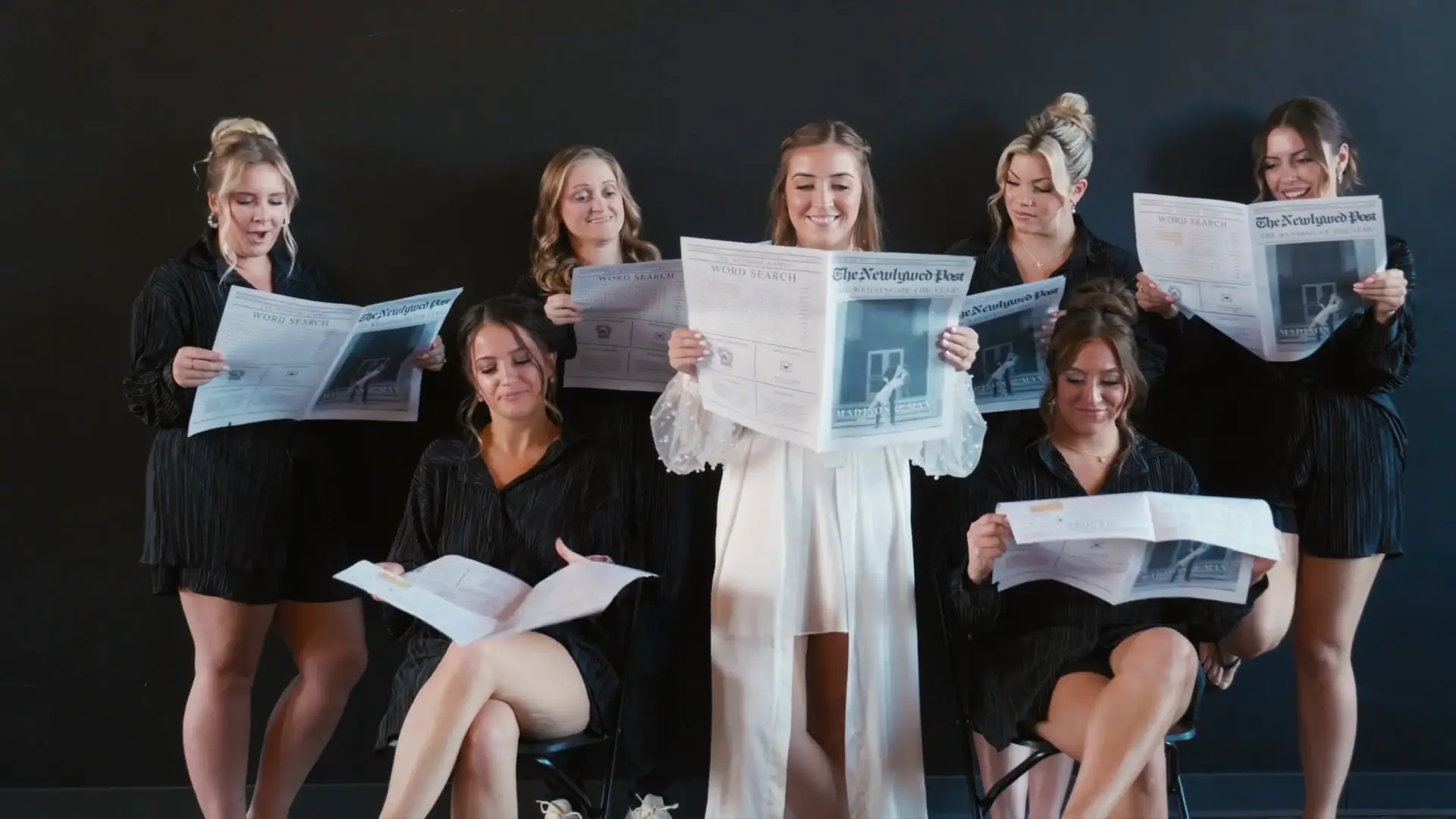 Madison and bridesmaids in matching black outfits, smiling and reading customized newspapers titled "The Newlywed Post" for Madison and Max's wedding. The joyful scene captures candid Michigan charm with a modern, playful twist.