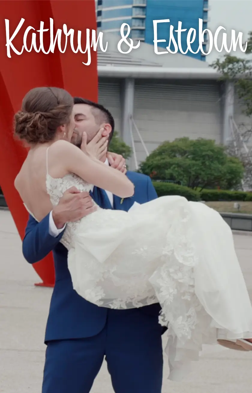 Kathryn and Esteban share a kiss in this snapshot from their wedding videography in Grand Rapids, Michigan. The groom lifts the bride in front of a vibrant red sculpture, capturing a joyful moment of their special day.