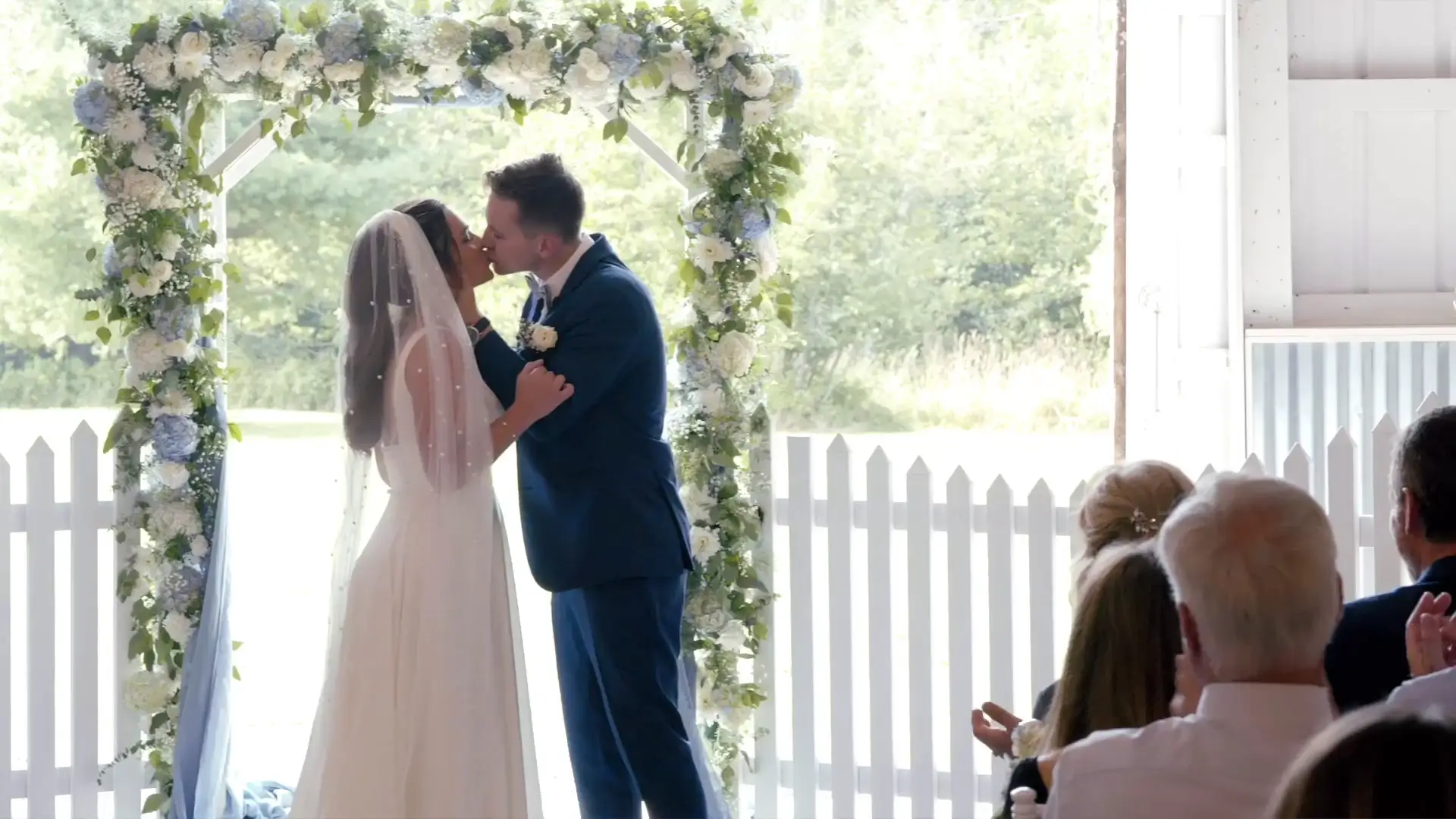 In this still from their wedding video, a bride and groom share a kiss under a floral arch during their ceremony, with guests seated nearby applauding.